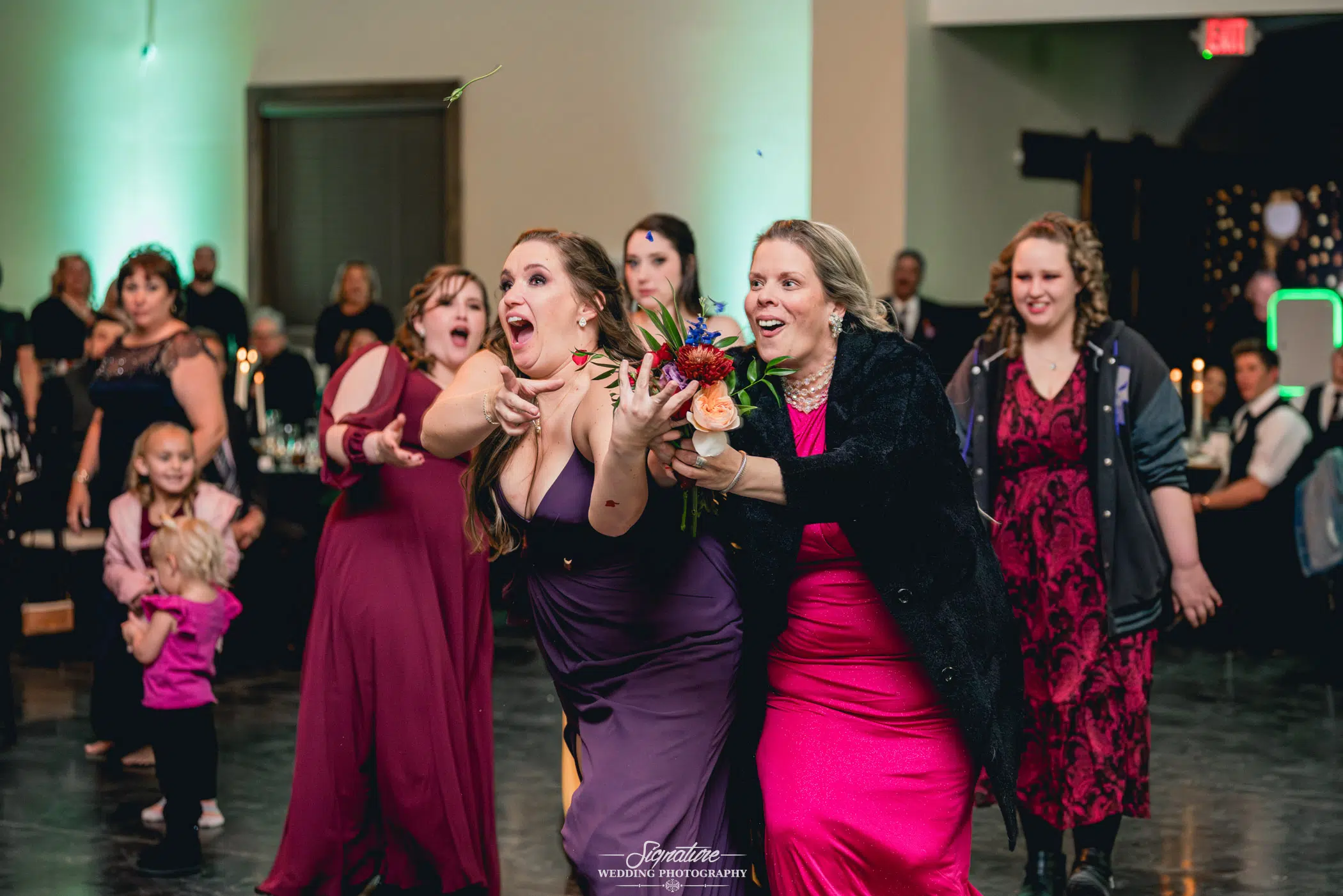 Image by Signature Wedding Photography A group of women enthusiastically reaching for a bouquet being tossed at a formal wedding event. They are in a festive setting, with onlookers in the background. The women are dressed in elegant outfits, and the scene captures a joyful, celebratory moment perfect for wedding photography.