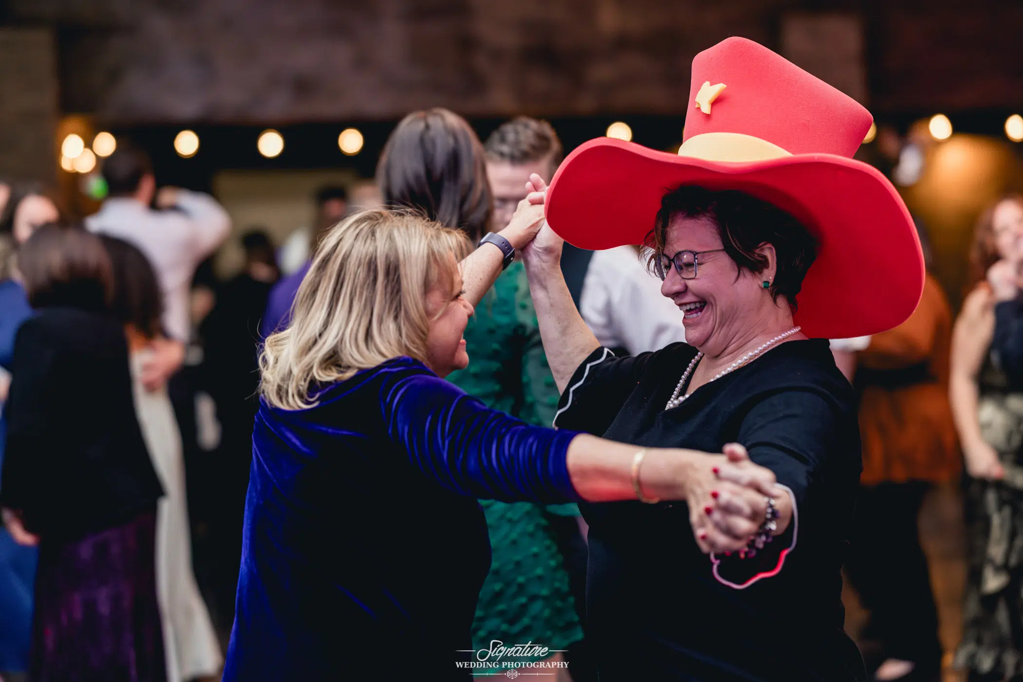 Image by Signature Wedding Photography Two women dance joyfully, one adorned with a large, bright red hat. They hold hands, smiling widely, captured in the art of wedding photography. The background is alive with others mingling in a festive indoor setting under warm lighting.