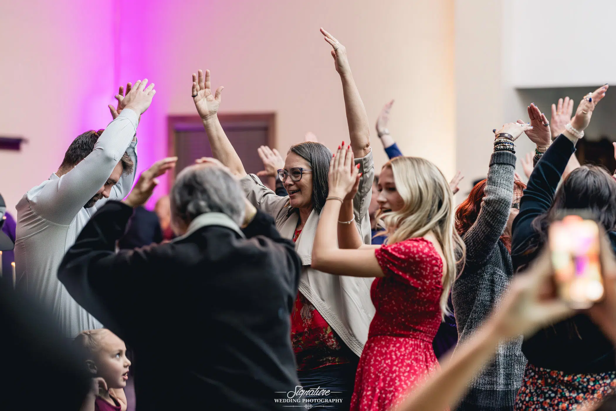 Image by Signature Wedding Photography People are dancing joyfully with their hands raised at a lively indoor event, perfectly capturing the essence of wedding photography. The group includes both men and women, with some dressed in formal attire. The atmosphere is festive, with warm lighting enhancing the celebratory mood.