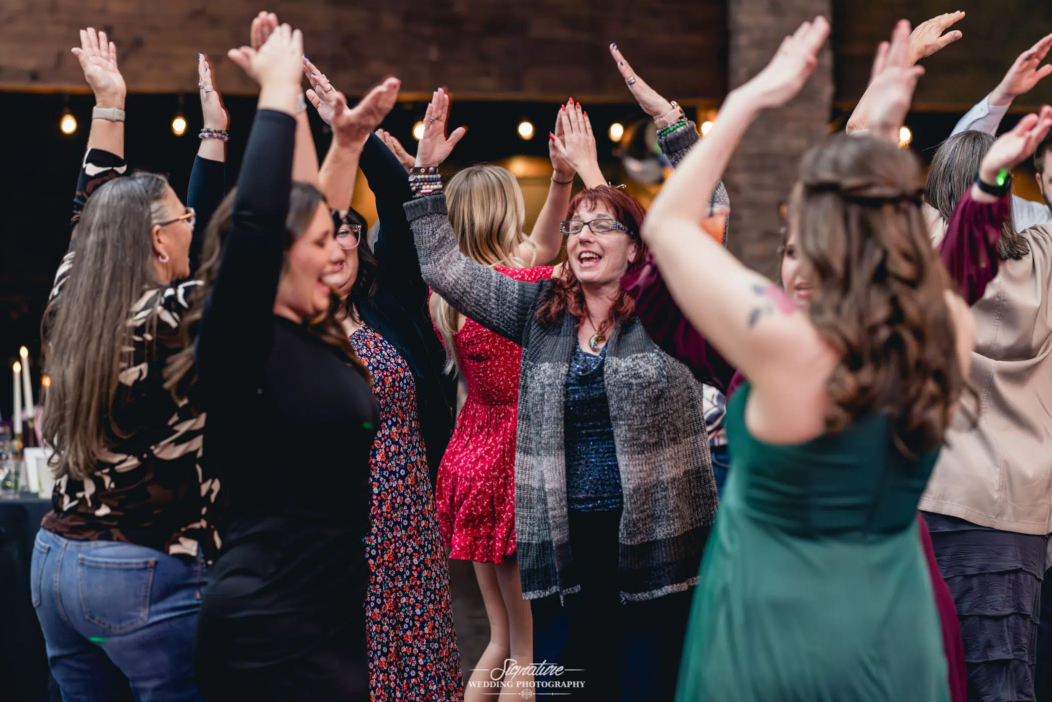 Image by Signature Wedding Photography A group of women with diverse outfits joyfully dance in a circle, their hands raised in celebration, as if captured by wedding photography at a festive indoor event. They smile amid the warm ambiance created by string lights adorning the background.