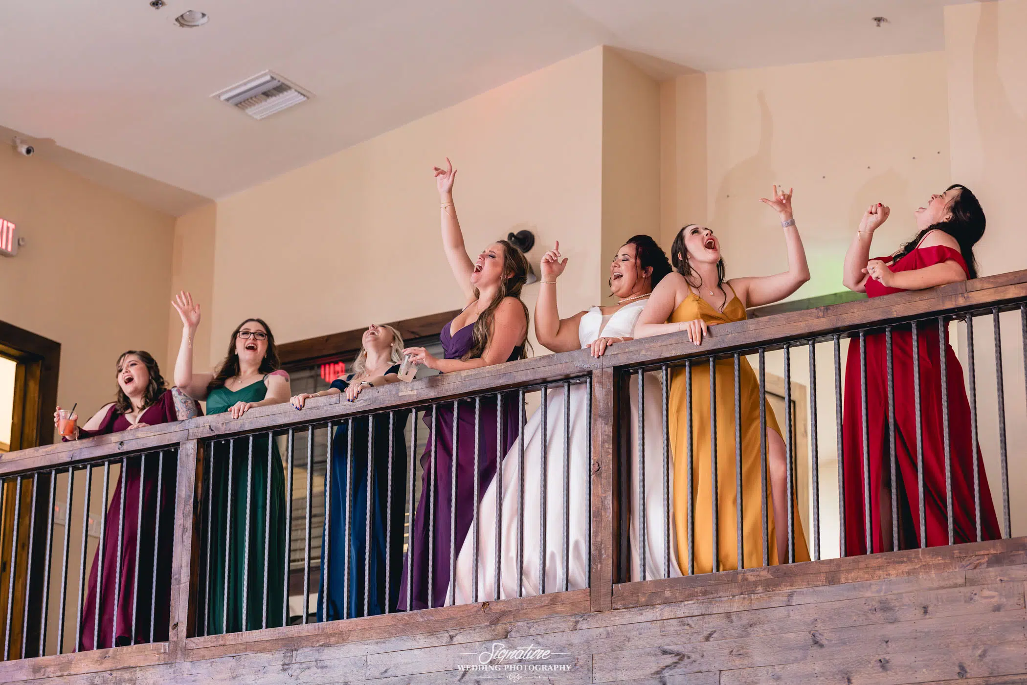 Image by Signature Wedding Photography A group of seven women stand on a wooden balcony indoors, exuding joy and enthusiasm as they cheer and point upwards. Clad in colorful dresses of purple, green, blue, white, mustard, and red, their vibrant energy perfectly captures the essence of wedding photography.