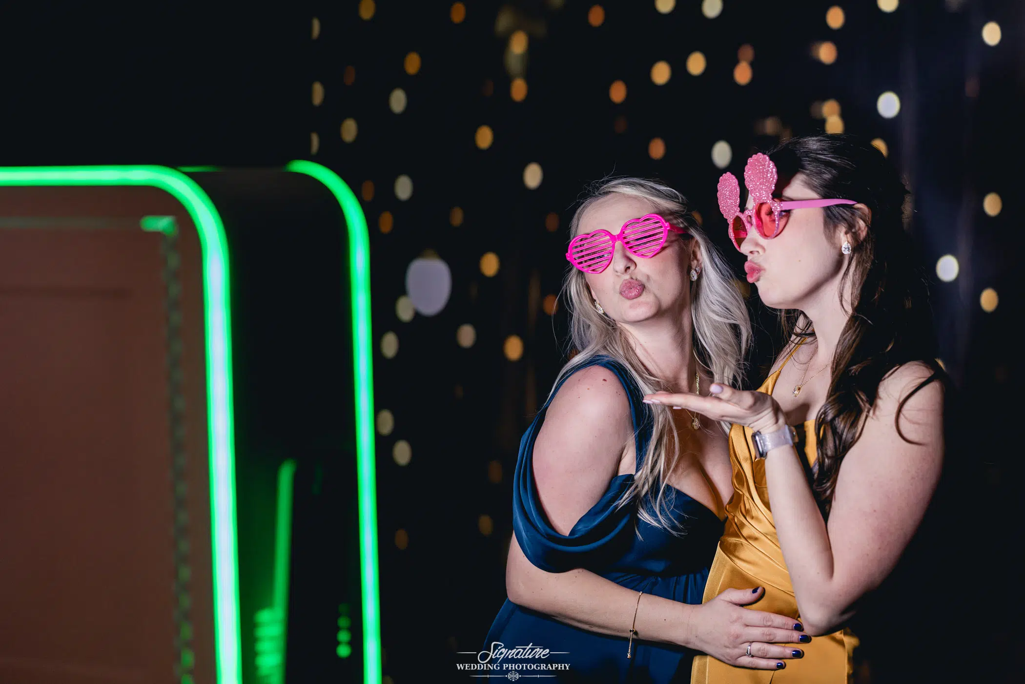 Image by Signature Wedding Photography Two women pose playfully, evoking the joy of wedding photography, in front of a dark background with bright dots. One wears a blue dress and heart-shaped glasses, the other a yellow dress with pink novelty sunglasses. They pout and blow kisses near a bright, green-lit booth to the left.