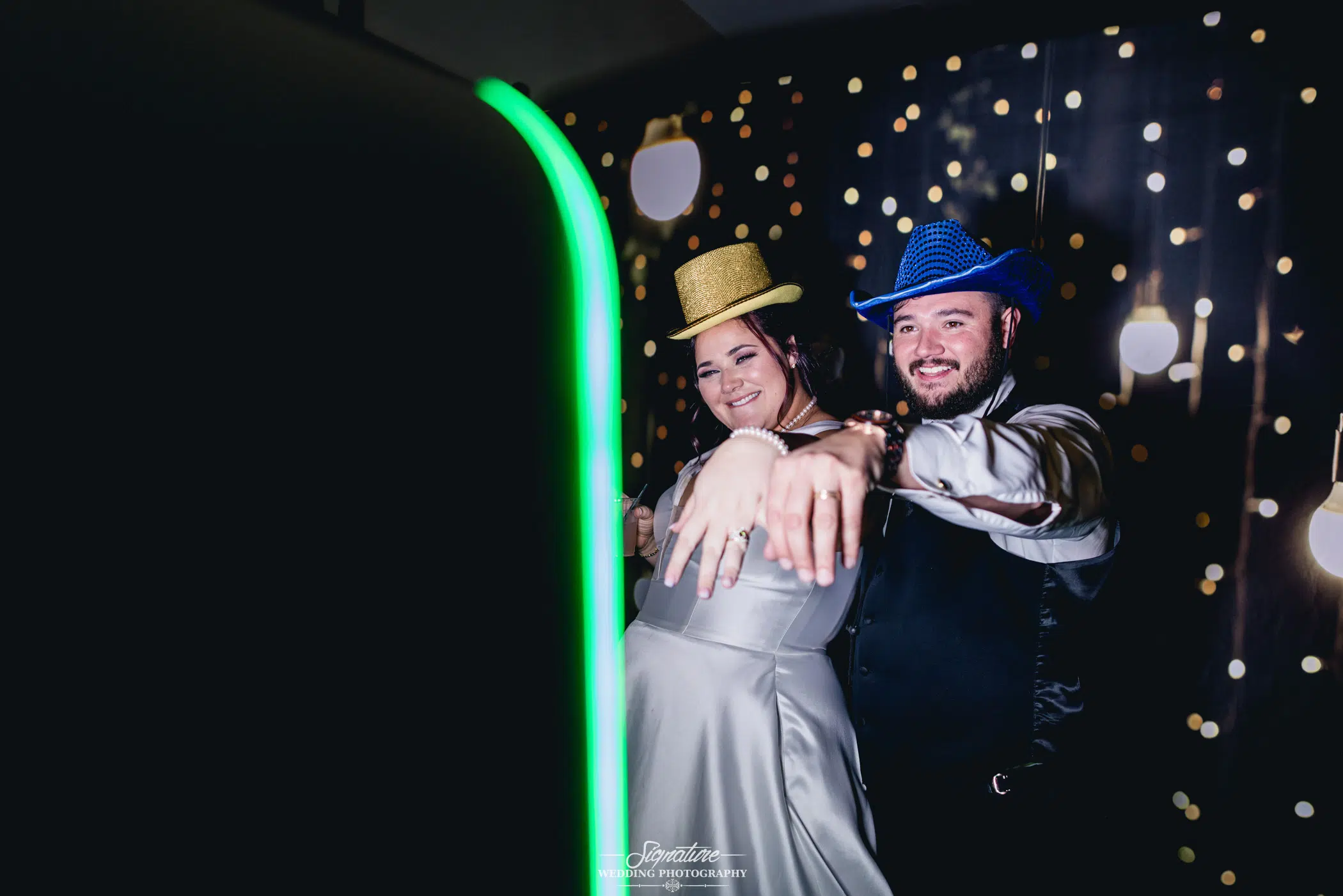 Image by Signature Wedding Photography A smiling couple poses in a photo booth, both wearing colorful hats. They extend their left hands to display rings, capturing a magical wedding photography moment. The background features a curtain with glowing string lights, creating a festive atmosphere.