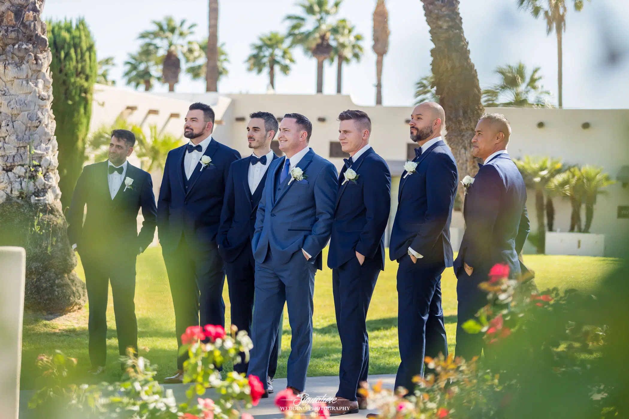 Image by Signature Wedding Photography Seven men in blue suits and bow ties stand together outdoors on a sunny day, surrounded by greenery and palm trees. They appear to be part of a wedding party, with most wearing white boutonnieres. Red flowers are visible in the foreground, perfectly capturing the essence of wedding photography.