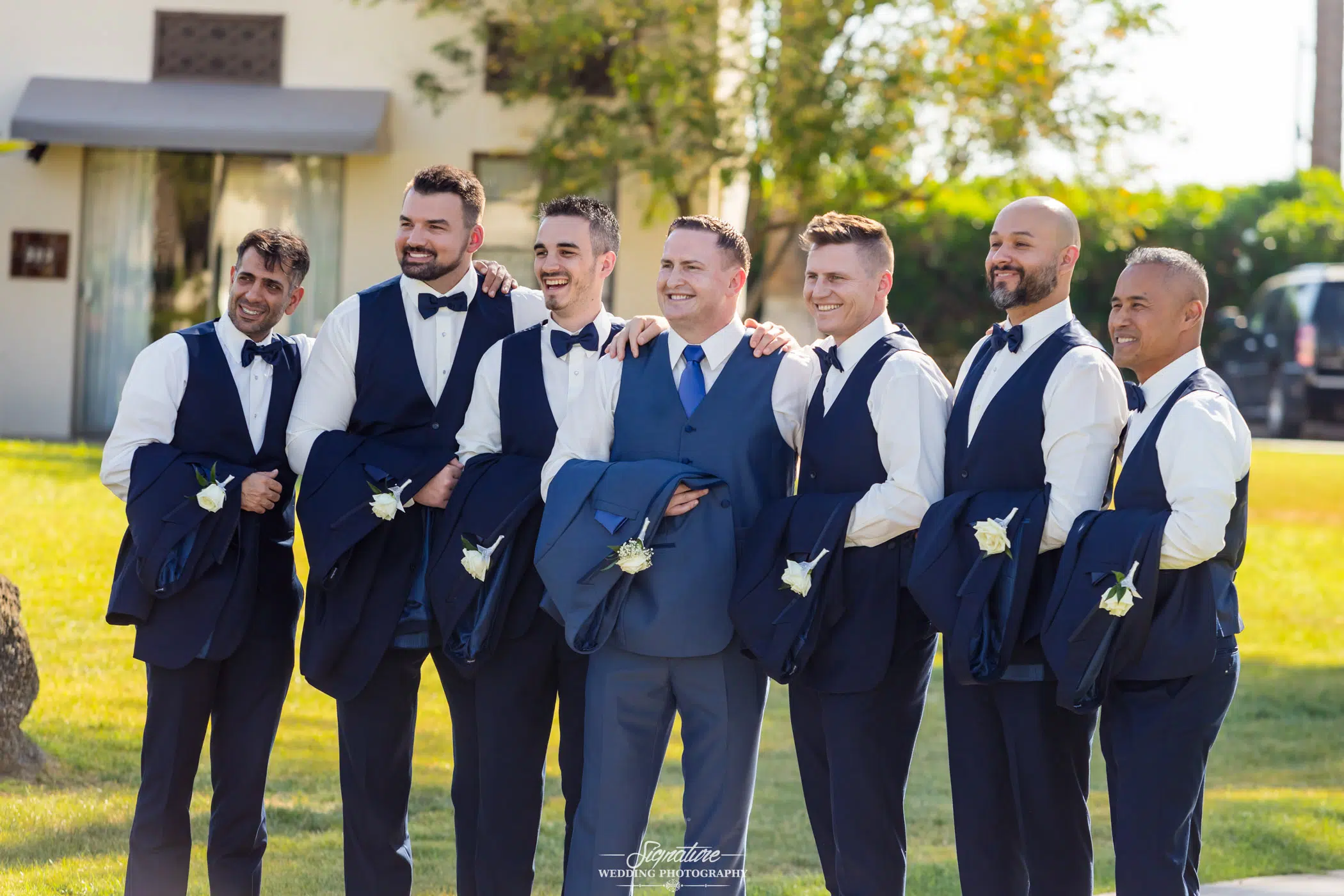 Image by Signature Wedding Photography Seven groomsmen in matching blue vests and bow ties stand outdoors, smiling and holding their jackets over their shoulders. Capturing the spirit of wedding photography, they are lined up on a sunny lawn with trees and a building in the background.
