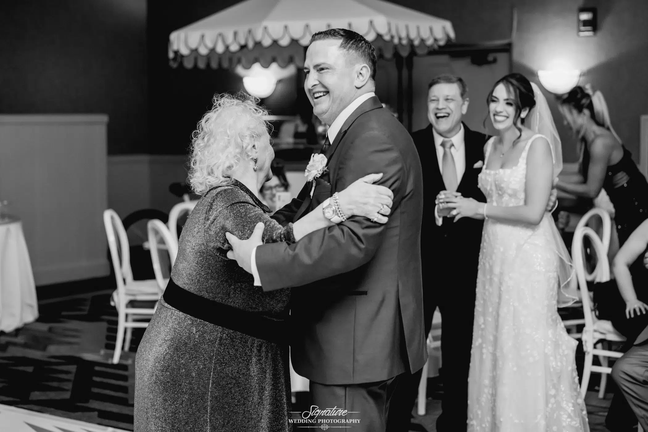Image by Signature Wedding Photography A black and white photo captures a joyful wedding photography moment at the reception. The groom dances with an elderly woman, both smiling radiantly. In the warmly lit room, the bride and guests laugh heartily in the background amidst tables and chairs.
