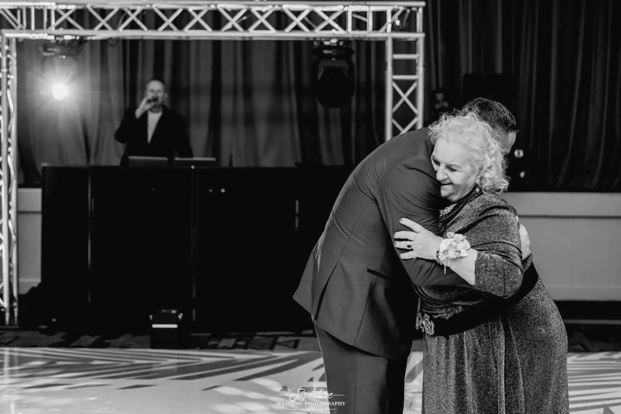 Image by Signature Wedding Photography A black and white wedding photography shot captures an elderly woman embracing a man on the dance floor. She wears a corsage, while a DJ stands nearby amidst audio equipment and lighting. A patterned floor and draped curtain add to the scene's nostalgic charm.