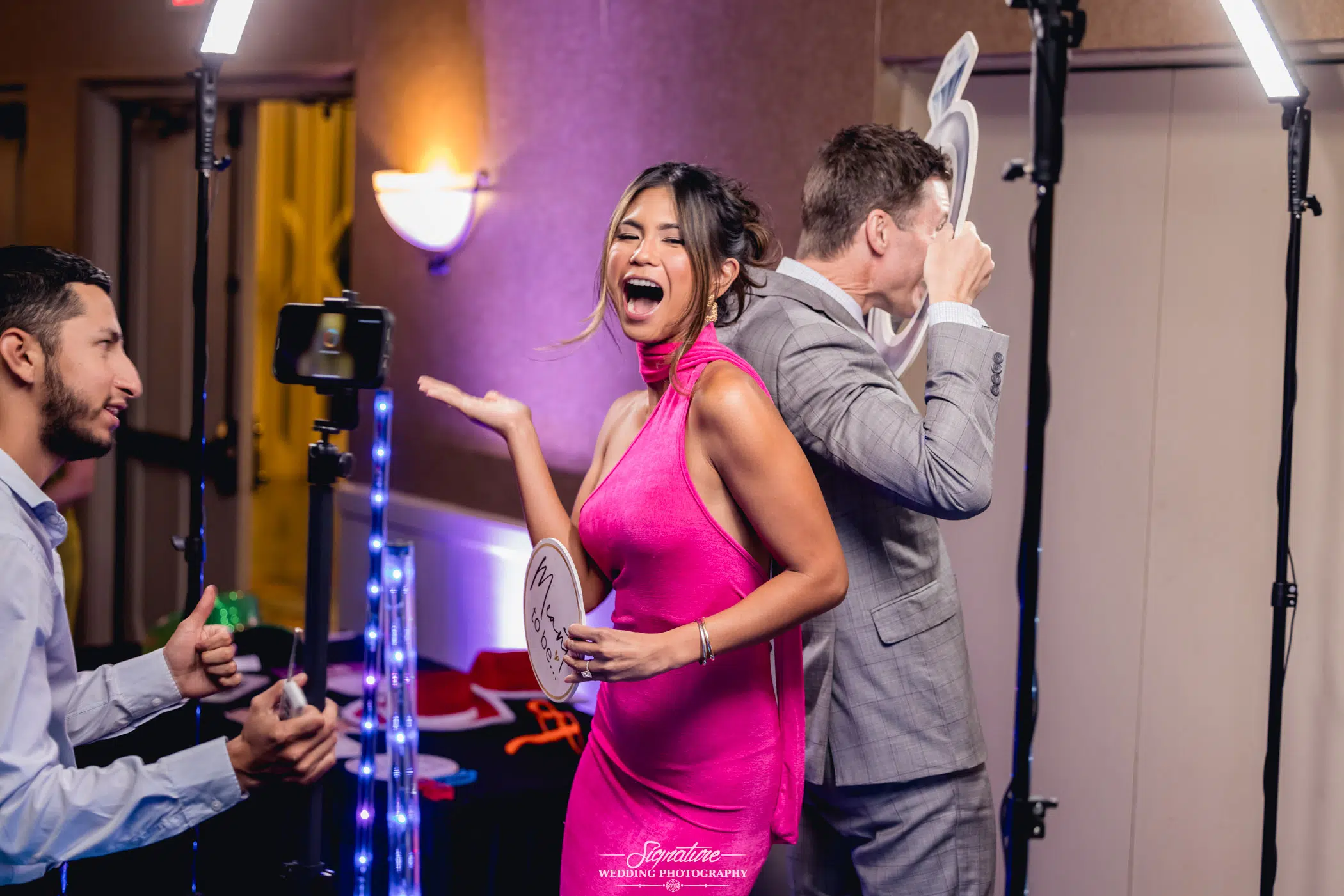 Image by Signature Wedding Photography A woman in a bright pink dress happily poses with a photo booth prop, while a man beside her holds another over his face. A wedding photographer captures the joyful moment as colorful lights illuminate the scene.