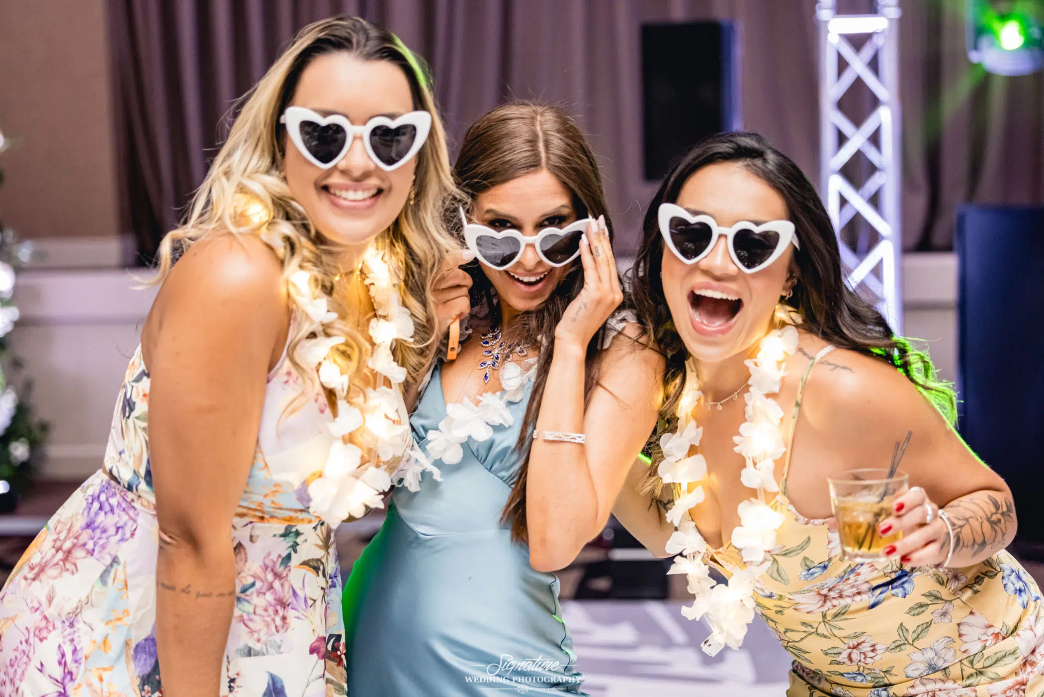 Image by Signature Wedding Photography Three women in floral dresses and heart-shaped sunglasses, smiling at a party. They are wearing flower garlands and holding drinks, capturing the festive joy of wedding photography.