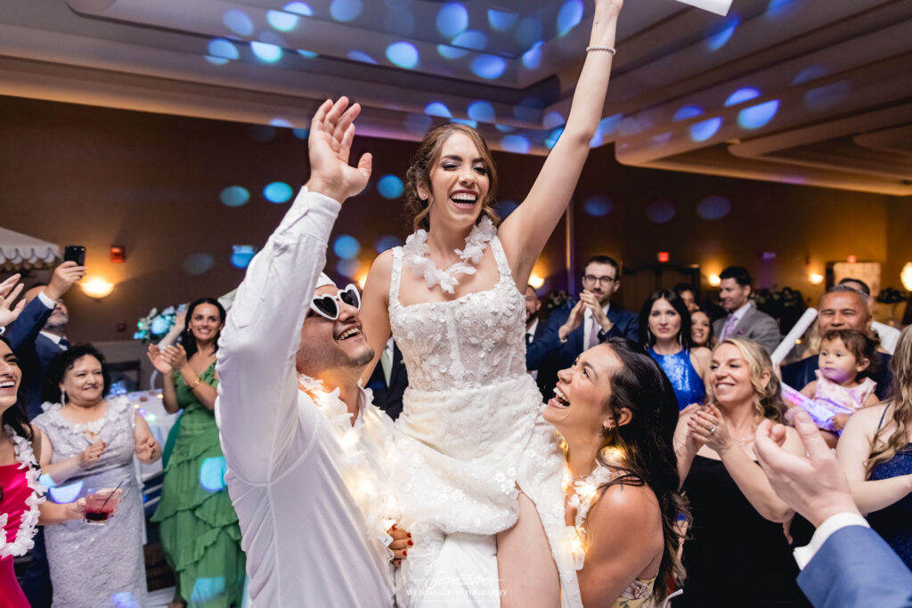 A joyful bride in a white dress is lifted up by two people at a lively wedding reception, captured beautifully in wedding photography. Guests around them smile and clap, some wearing leis, as dim lighting and colorful dots dance on the walls.