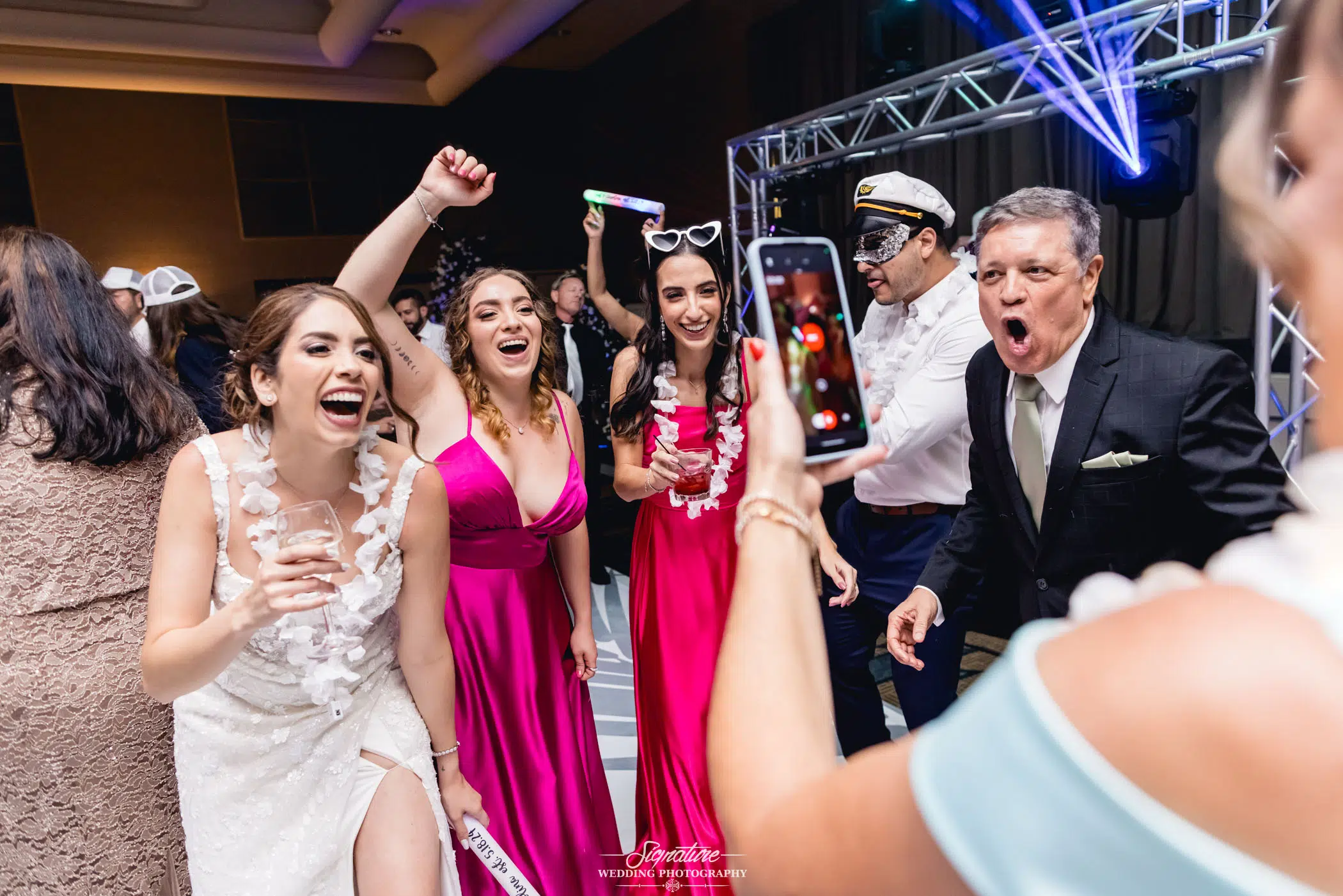 Image by Signature Wedding Photography A group of people dressed in formal attire are joyfully dancing and singing at what appears to be a wedding party. The women in vibrant dresses and a man in a suit are captured mid-celebration, with a woman in white holding her smartphone. Bright lights illuminate the scene, perfect for wedding photography.