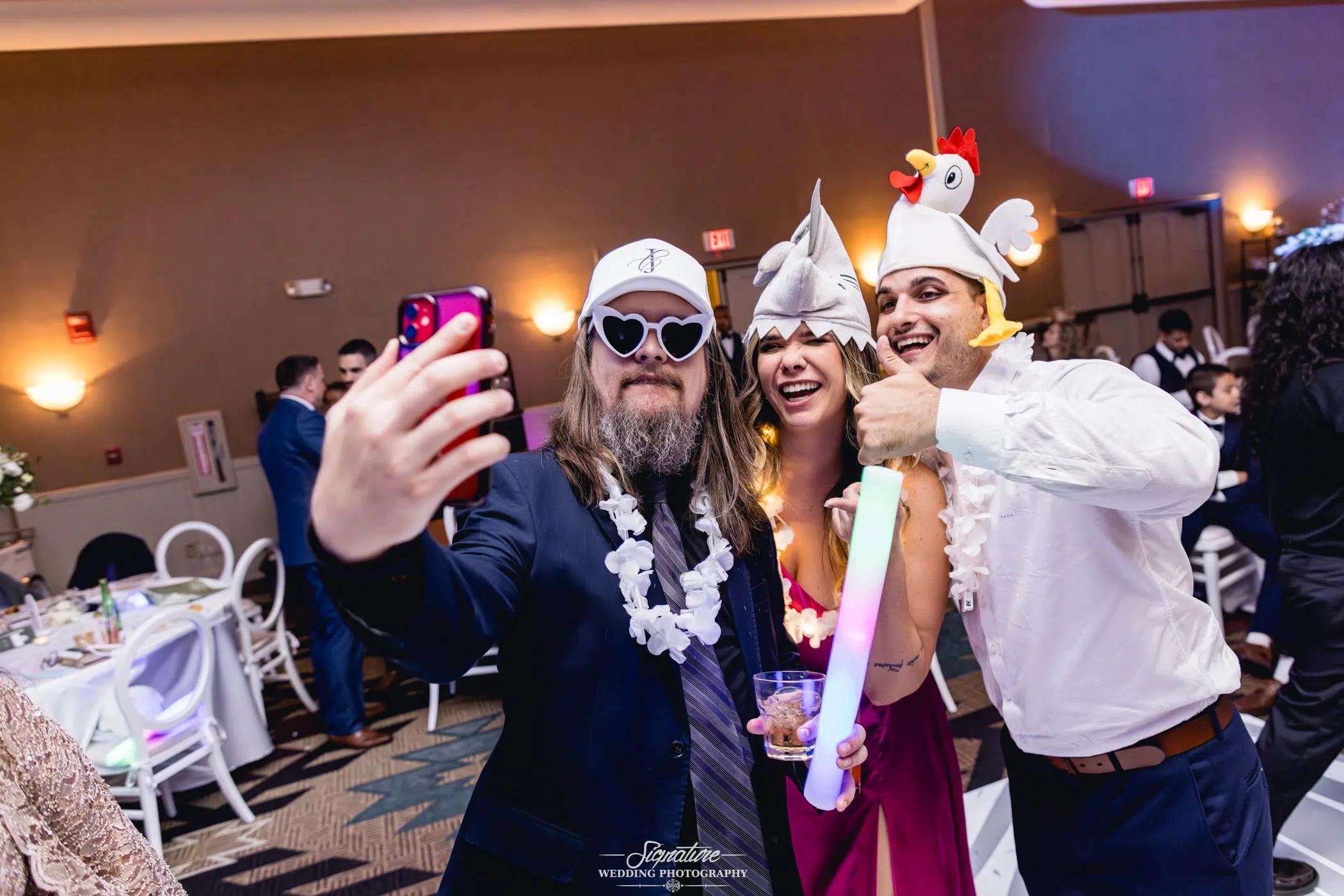 Image by Signature Wedding Photography Three people in festive attire take a selfie at an indoor celebration. One sports heart-shaped sunglasses and a Viking hat, another a shark hat, and the third a chicken hat. With radiant smiles, they capture the joy akin to candid wedding photography moments.