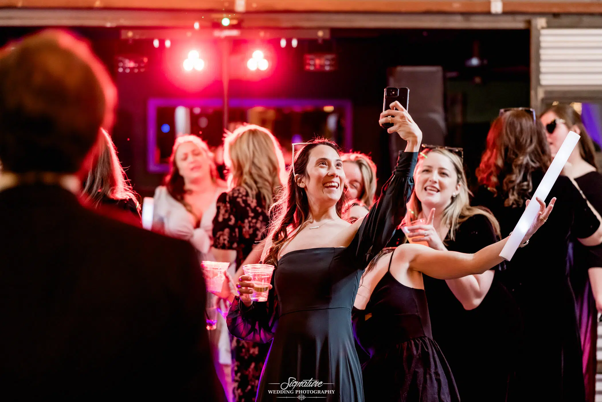 Image by Signature Wedding Photography At an indoor event illuminated by red lighting and DJ equipment, a group of people, mostly women in dresses, dance joyfully. Capturing the moment like a scene from wedding photography, one woman in a dark dress takes a selfie. Everyone appears to be having fun.