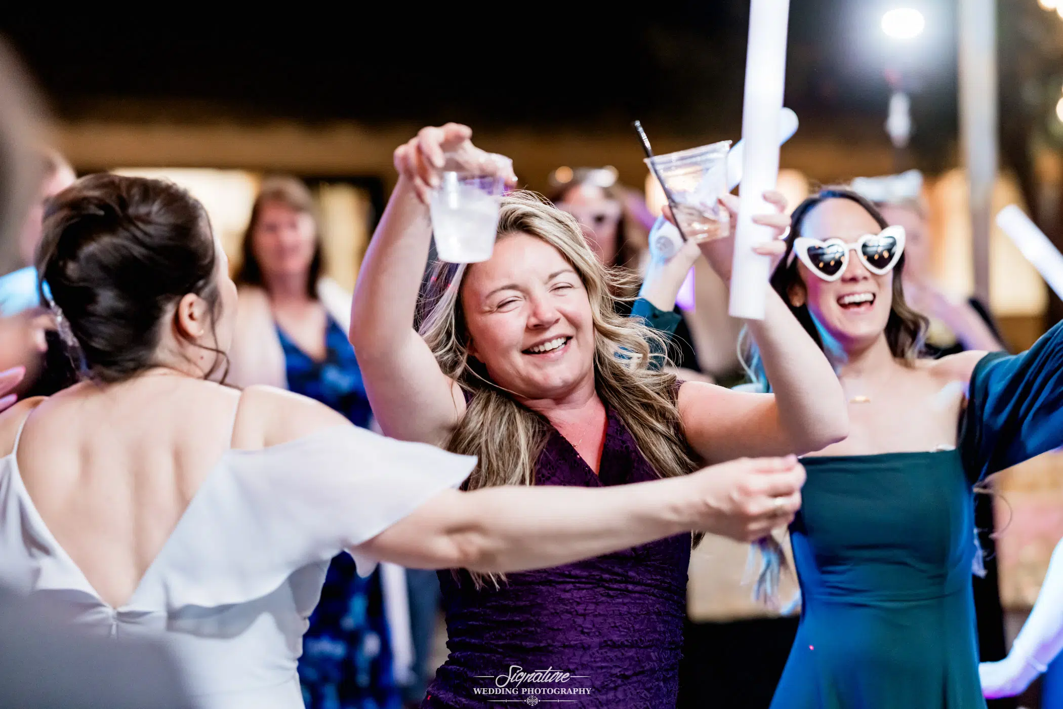 Image by Signature Wedding Photography A joyful woman in a purple dress dances amidst the wedding photography, holding a drink in one hand. She smiles widely, while a woman beside her sports heart-shaped sunglasses. The scene is lively, with guests and colorful lights filling the background.