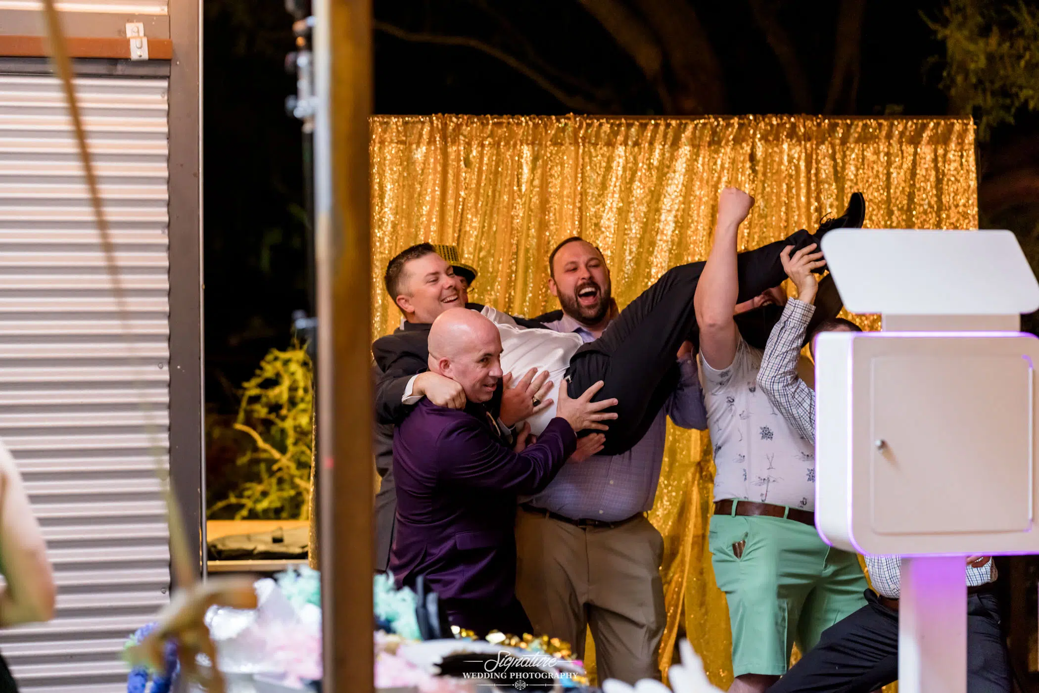 Image by Signature Wedding Photography A group of men are joyfully lifting another man in the air at a wedding event. Standing in front of a golden sequin curtain, the scene captures the essence of wedding photography with its lively and celebratory atmosphere, complete with assorted decorations.