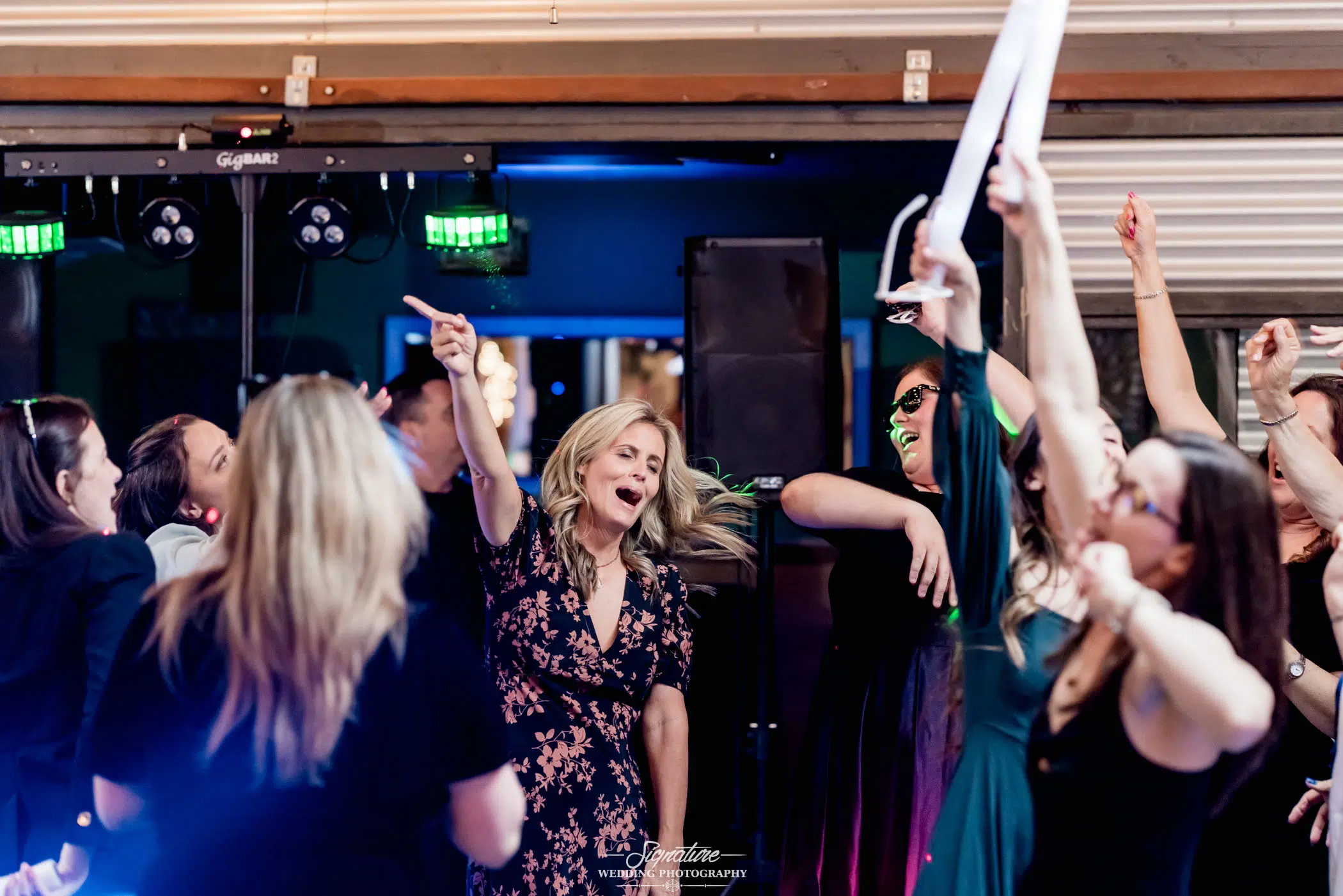 Image by Signature Wedding Photography A group of people energetically dancing at a wedding party. A woman in the foreground points upward, surrounded by others raising their arms and smiling. There's colorful lighting, a DJ booth in the background, and a photographer capturing every joyful moment of this vibrant wedding celebration.