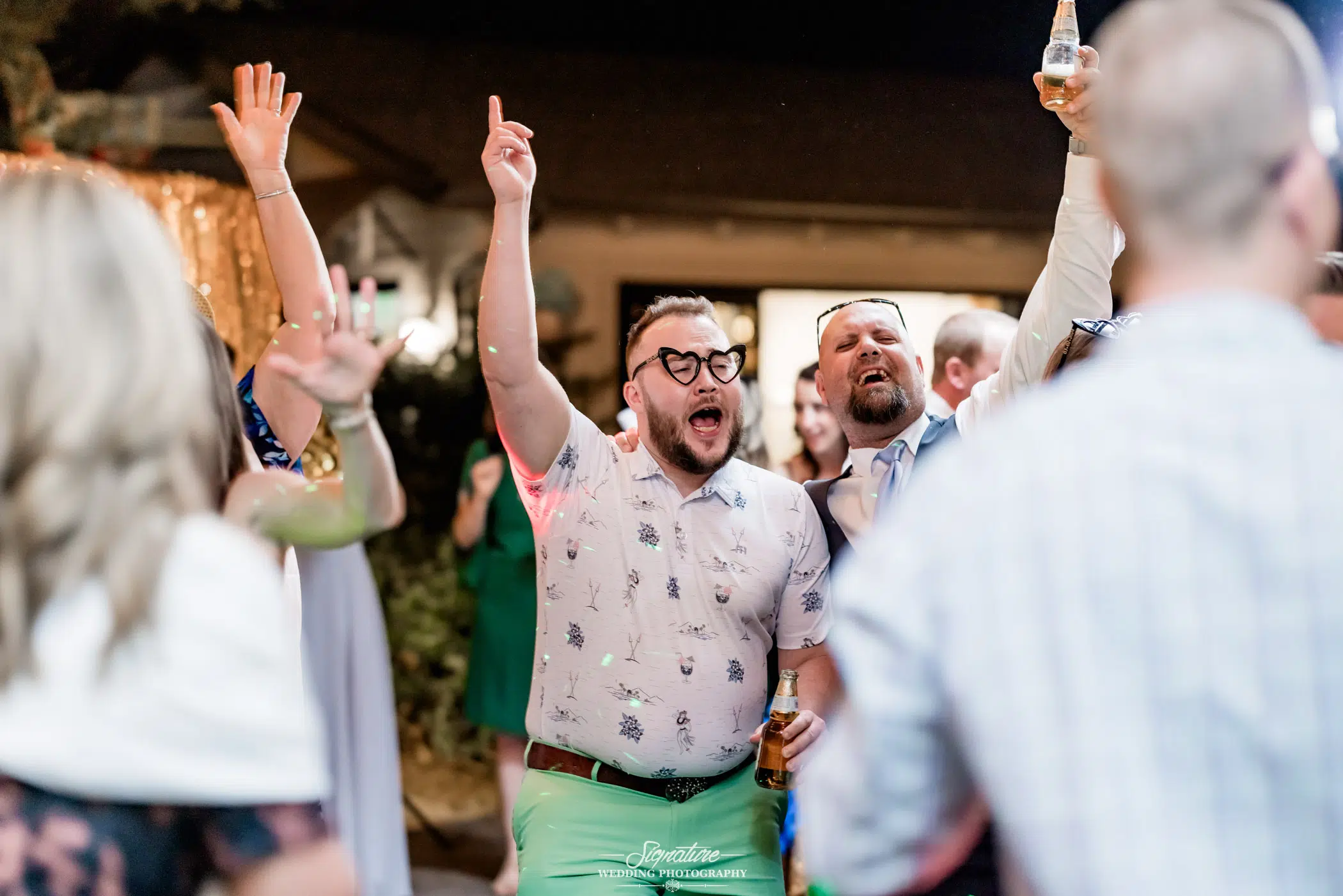 Image by Signature Wedding Photography Group of people dancing and singing at an outdoor event. A man in a floral shirt and glasses raises his hand enthusiastically, holding a drink. Captured moments like these are the essence of wedding photography, as others around him revel in the lively atmosphere.