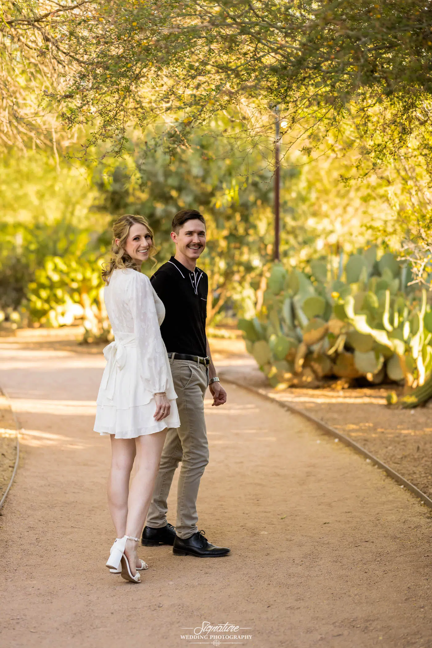 Image by Signature Wedding Photography A couple strolls hand in hand on a sunlit dirt path, surrounded by lush greenery and cacti. The woman, in a white dress and heels, and the man, in a black shirt and beige pants, both smile at the camera. This moment beautifully captures the essence of wedding photography.