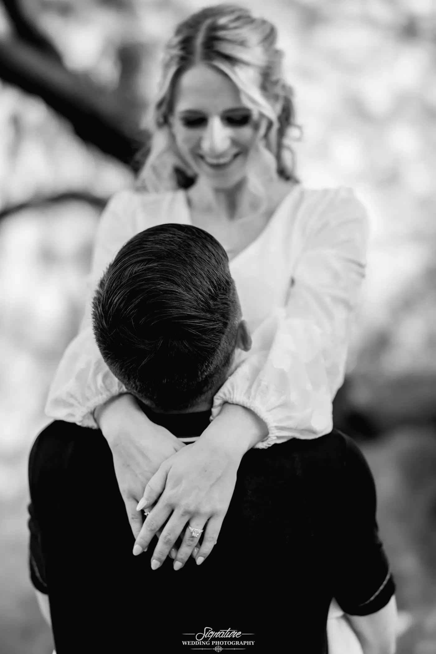 Image by Signature Wedding Photography A black and white photo captures a woman in a white dress, embodying the essence of wedding photography, smiling down at a man as she wraps her arms around his neck. The man faces away from the camera. Both are outdoors, with blurred trees in the background.