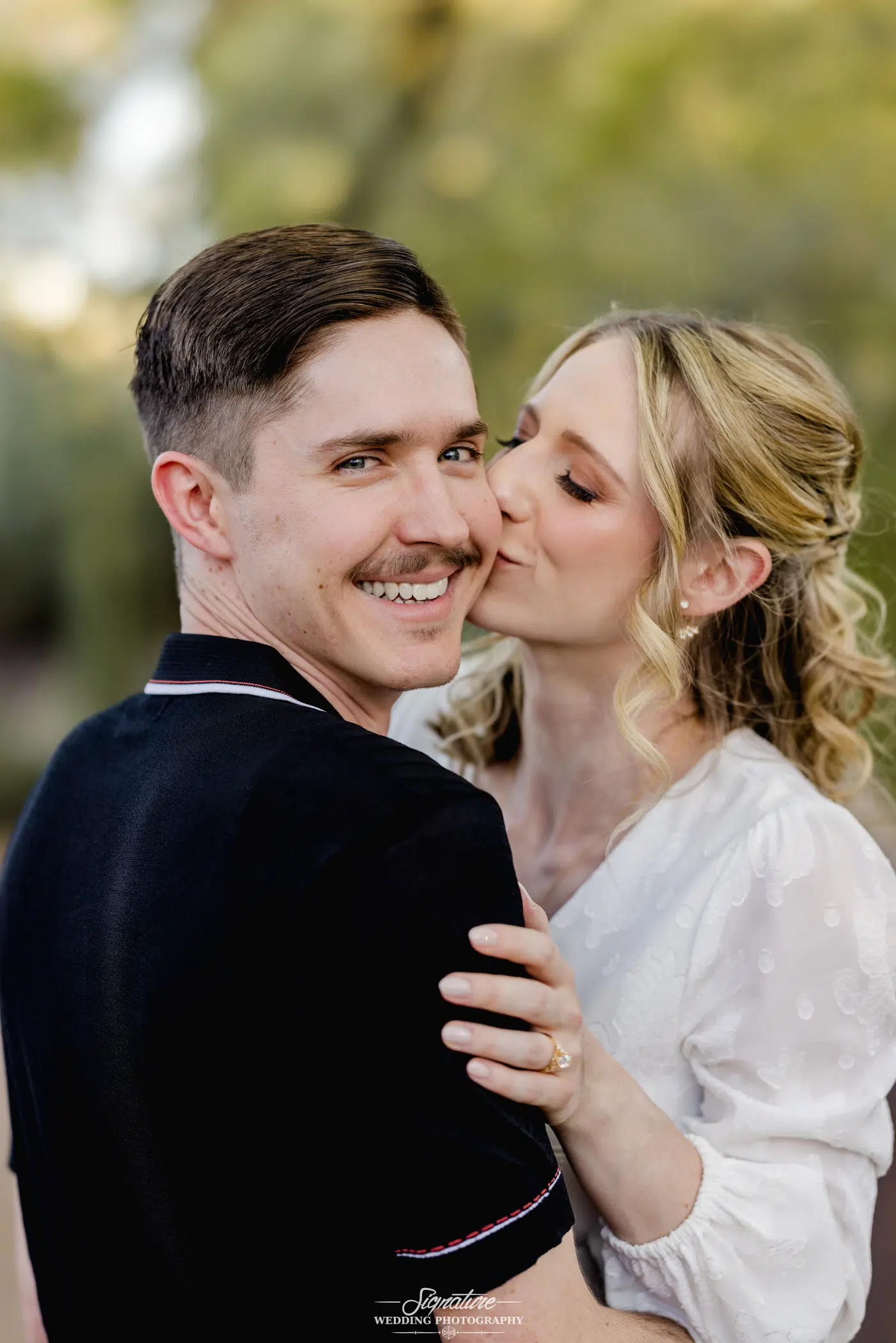 Image by Signature Wedding Photography A woman with curly blonde hair, wearing a white blouse, kisses a man with short brown hair and a mustache on the cheek. The man, in a black shirt, smiles at the camera. Captured outdoors against a blurred green background, it exudes the joy of candid wedding photography.