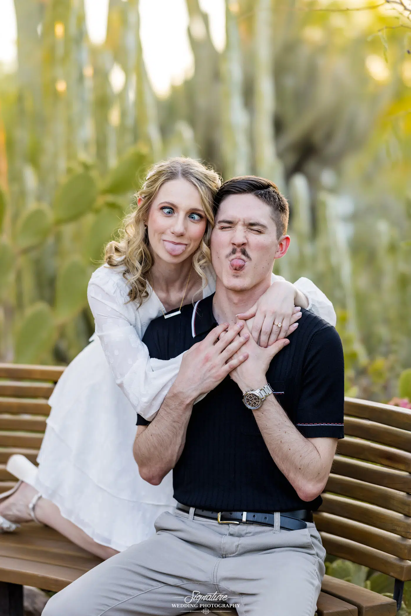 Image by Signature Wedding Photography A couple playfully poses on a wooden bench surrounded by cacti, embodying a whimsical take on wedding photography. The woman in a white dress wraps her arms around the man's shoulders, both sticking their tongues out. In a black shirt and light pants, he looks amused.