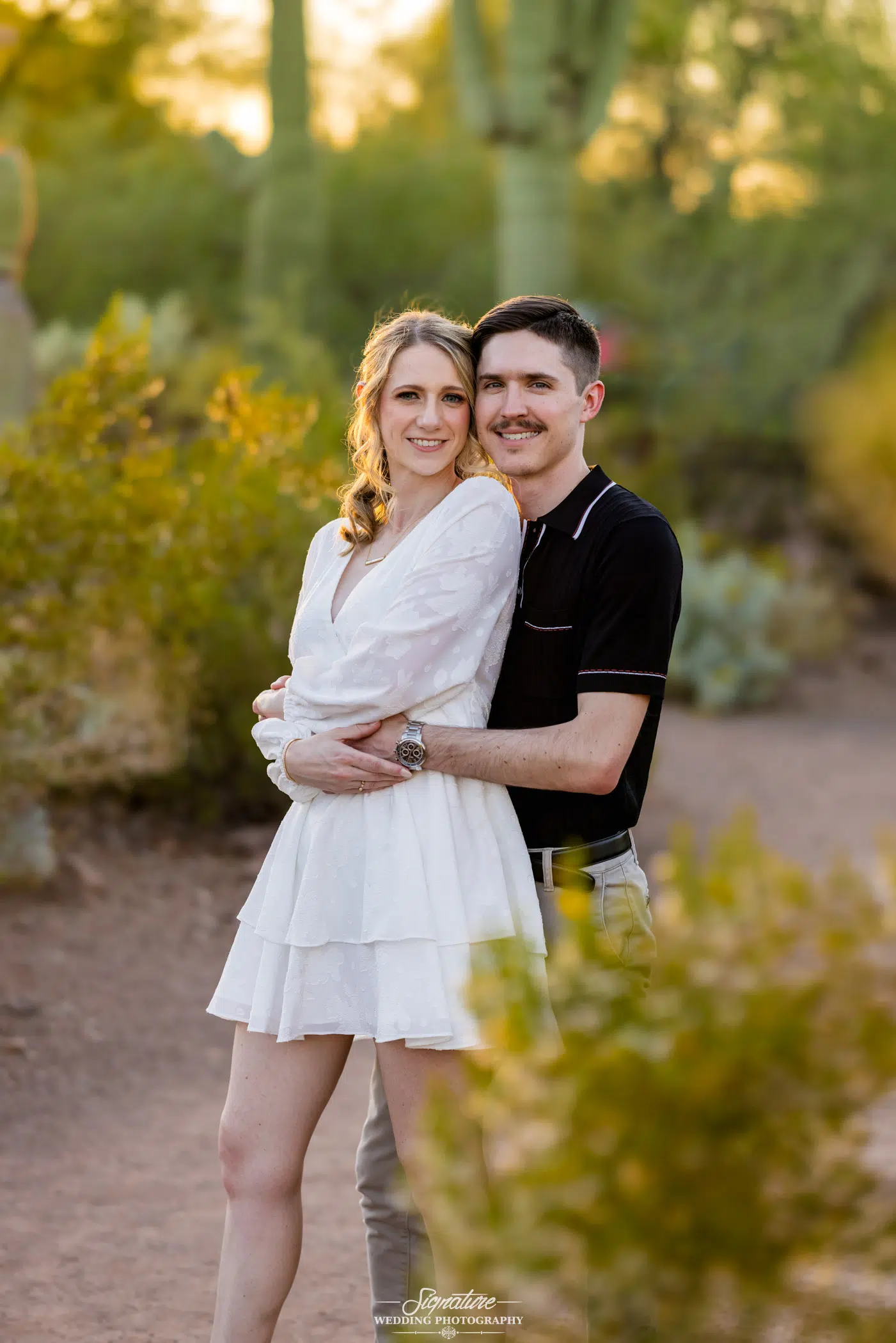 Image by Signature Wedding Photography A smiling couple stands on a desert path surrounded by cacti and bushes. The woman in a white dress is in front, while the man in a black shirt and khaki pants hugs her from behind—it’s the perfect capture for wedding photography as sunlight filters through the greenery.