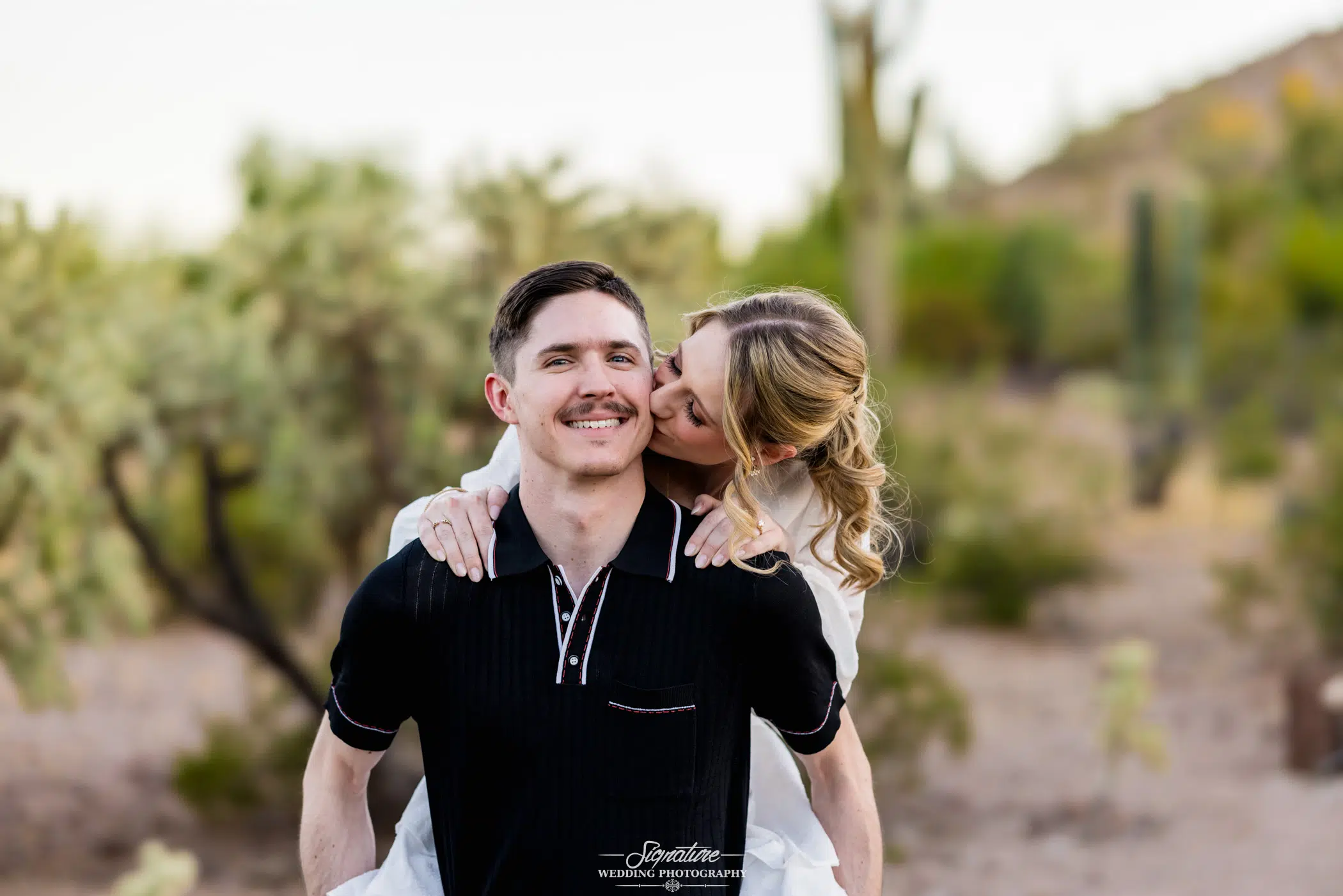 bride to be, kissing her husband to be on the cheek with an Arizona desert background