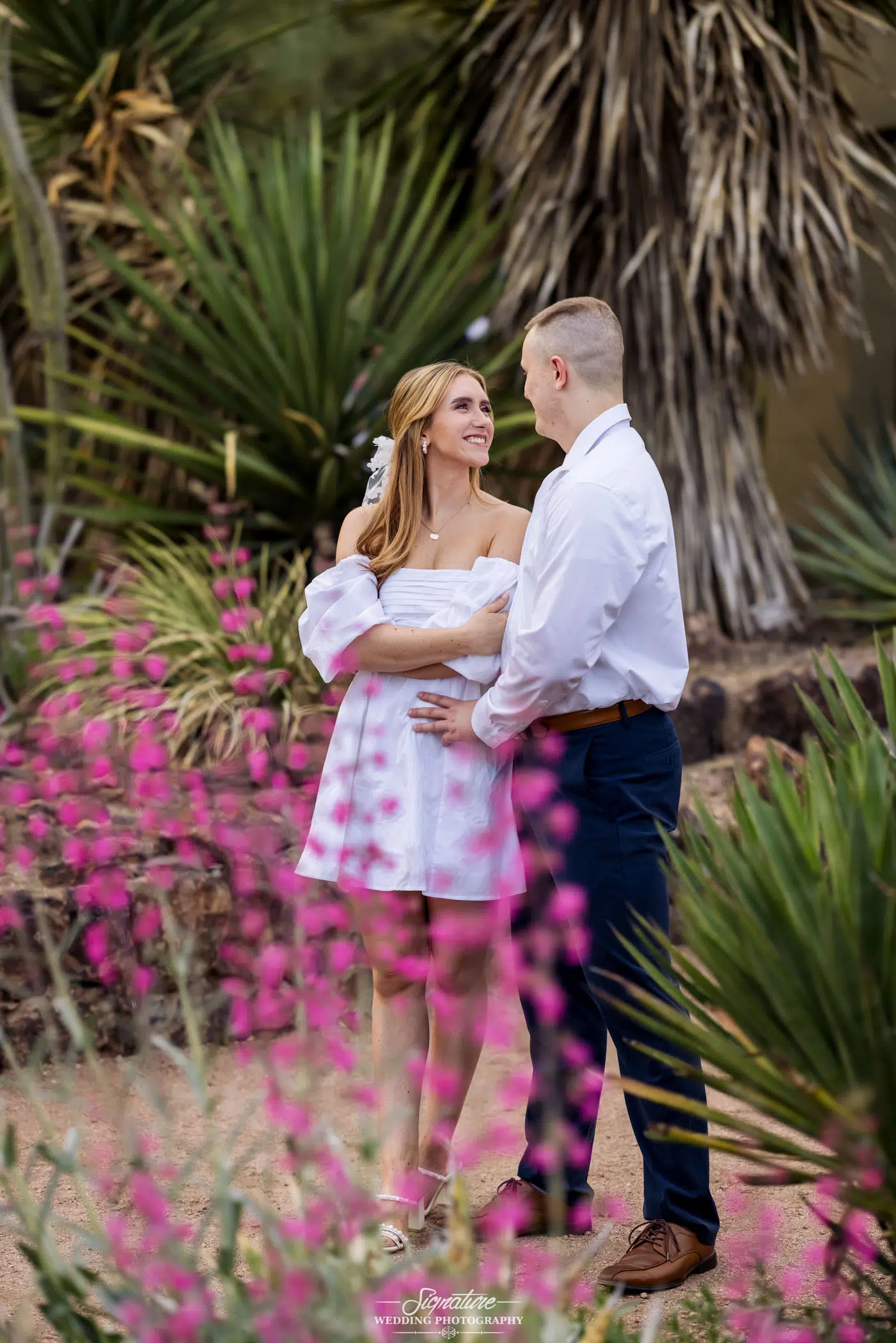 Photo by Tavits Photography In a picturesque garden adorned with pink flowers and lush green foliage, a couple stands as if in a scene from wedding photography. The woman, dressed in white, gazes up smiling at her partner in a white shirt and dark pants. They embrace tenderly amidst the natural beauty.