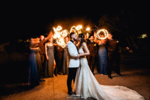 A bride and groom are kissing while a group of friend behind them wave sparklers for an artistic effect