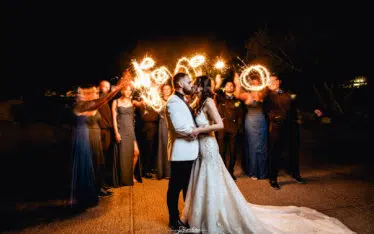 A bride and groom are kissing while a group of friend behind them wave sparklers for an artistic effect