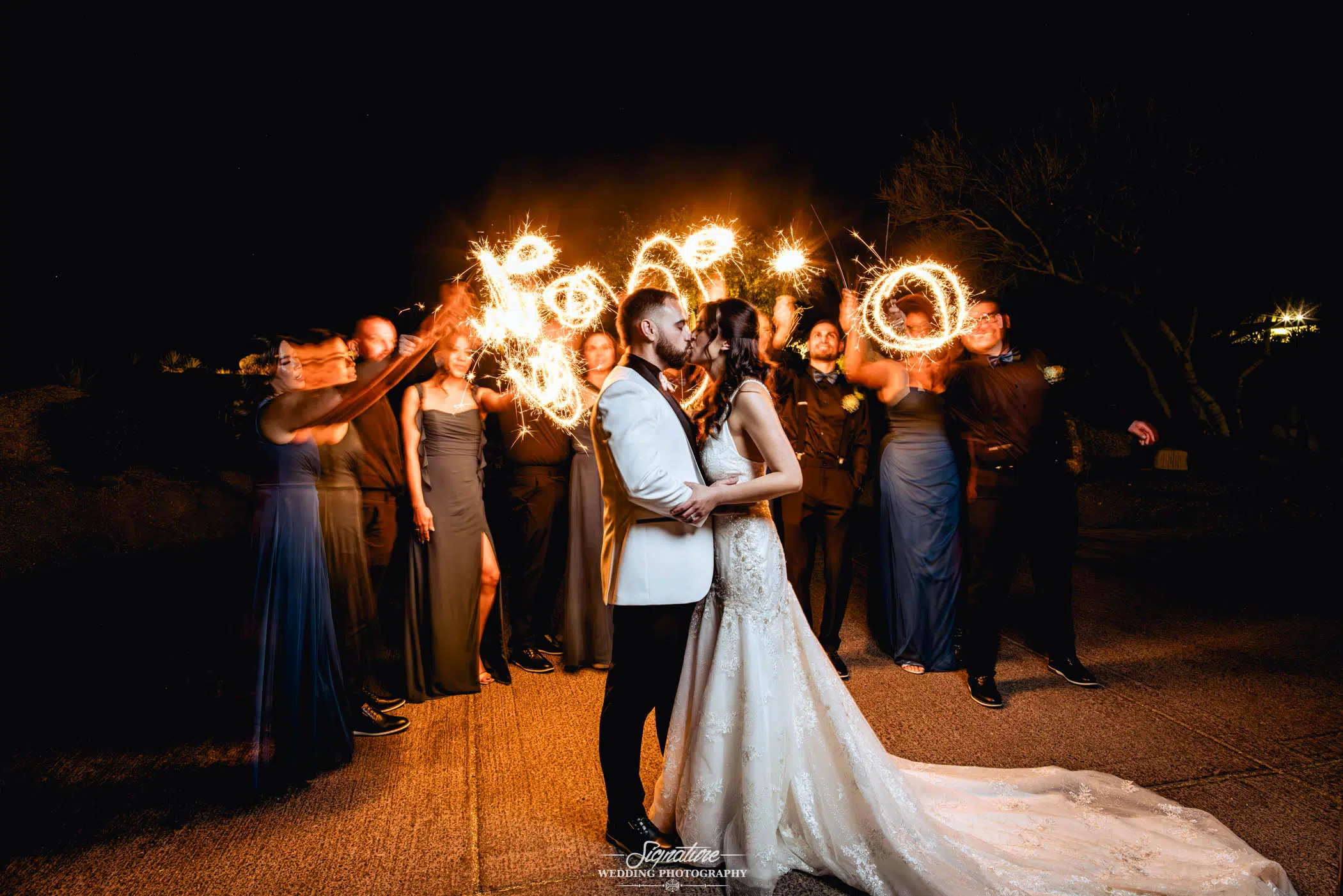 A bride and groom are kissing while a group of friend behind them wave sparklers for an artistic effect