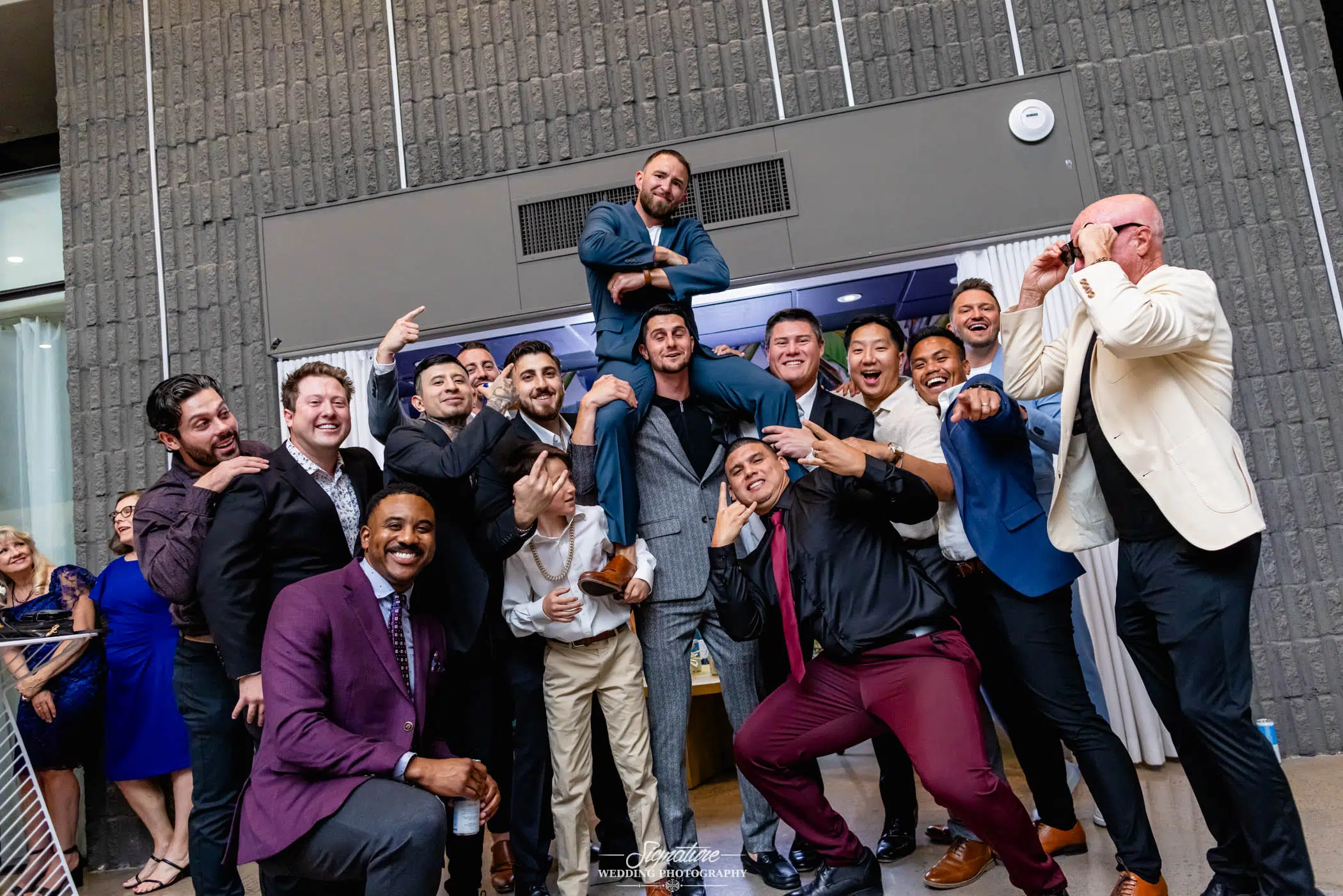 Photo by Tavits Photography A group of men dressed in suits pose playfully at an indoor event, with one man lifted on the shoulders of others while the rest strike fun, silly poses and smile. The background features a textured gray wall.