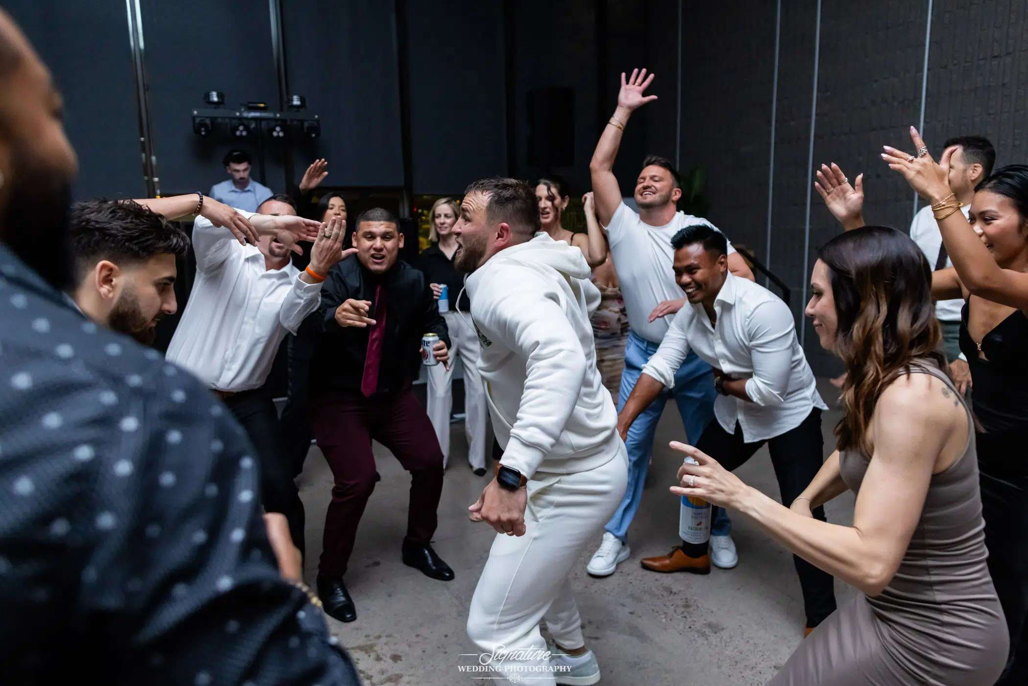 Photo by Tavits Photography A group of people enthusiastically dancing in a circle at an indoor event, with a man in a white outfit in the center, everyone smiling and enjoying the celebration.