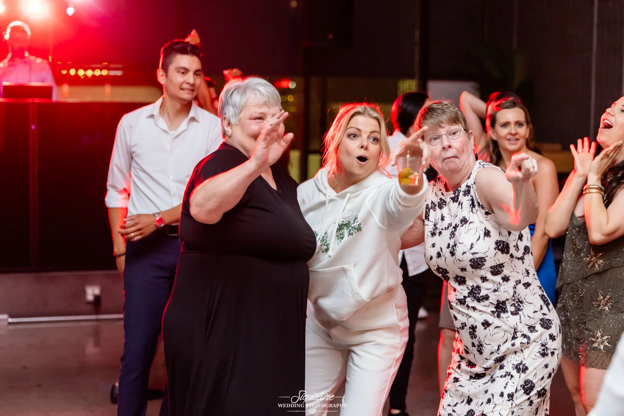 Photo by Tavits Photography A group of people dancing and smiling at a lively party, with three women in front enthusiastically pointing toward the camera. A DJ plays in the background under red lighting.