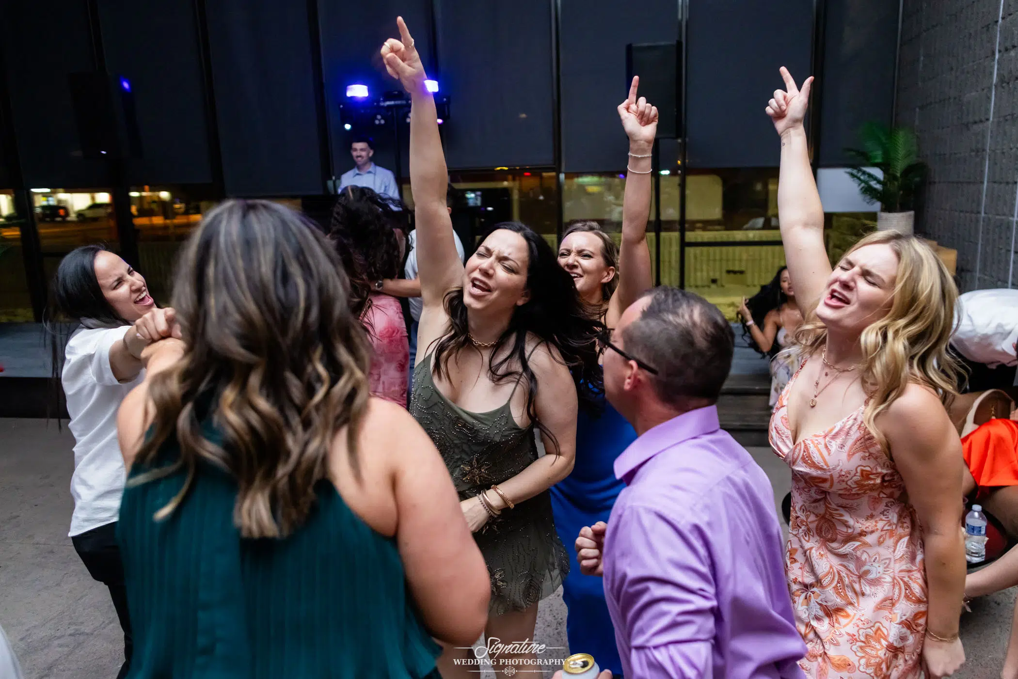 Photo by Tavits Photography A group of people joyfully dance with their hands raised at a party or wedding reception, smiling and celebrating together under bright lights.