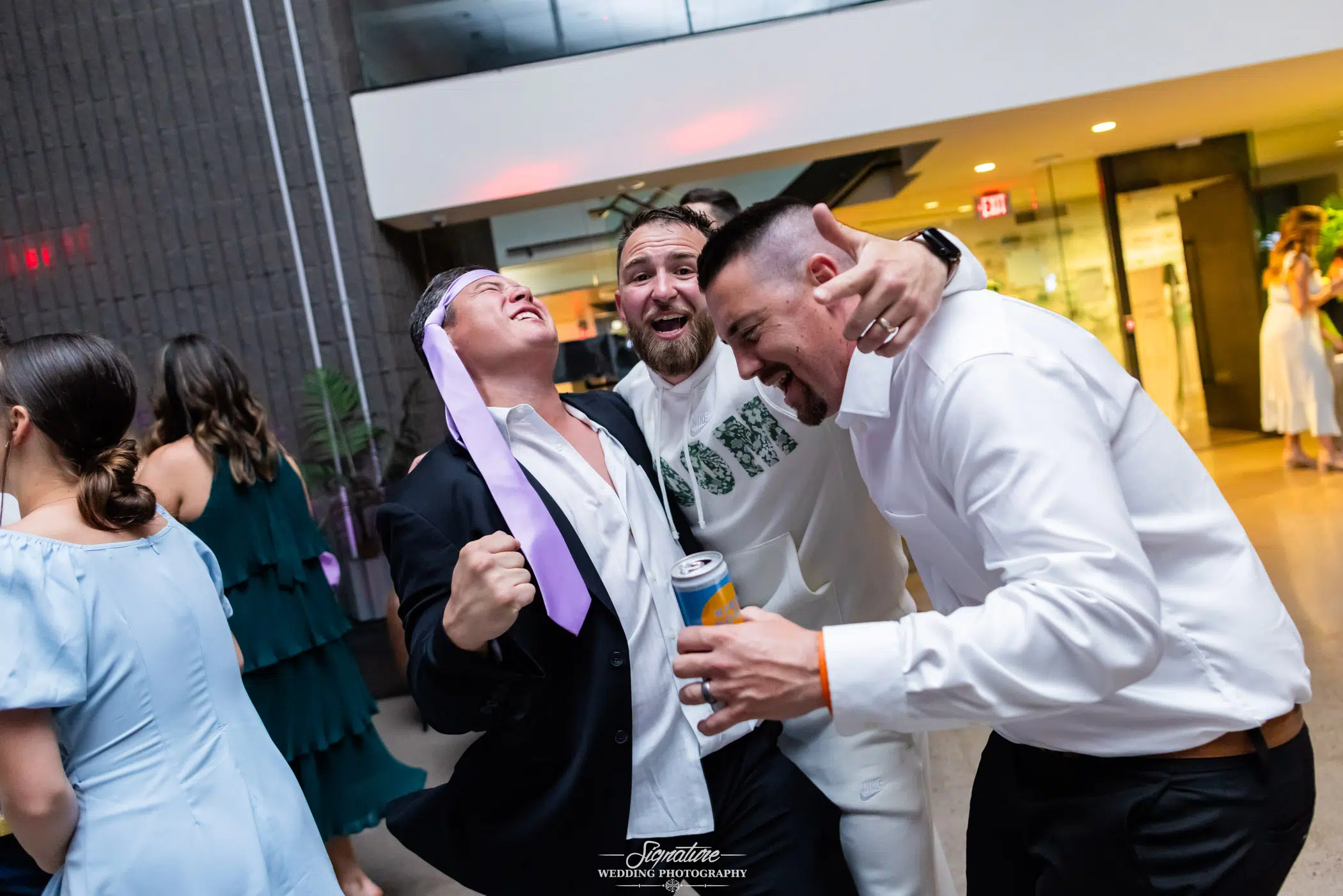 Photo by Tavits Photography Three men in dress shirts laugh and celebrate together at a lively indoor event; one man has a purple tie wrapped around his head, and another holds a can, while people mingle in the background.