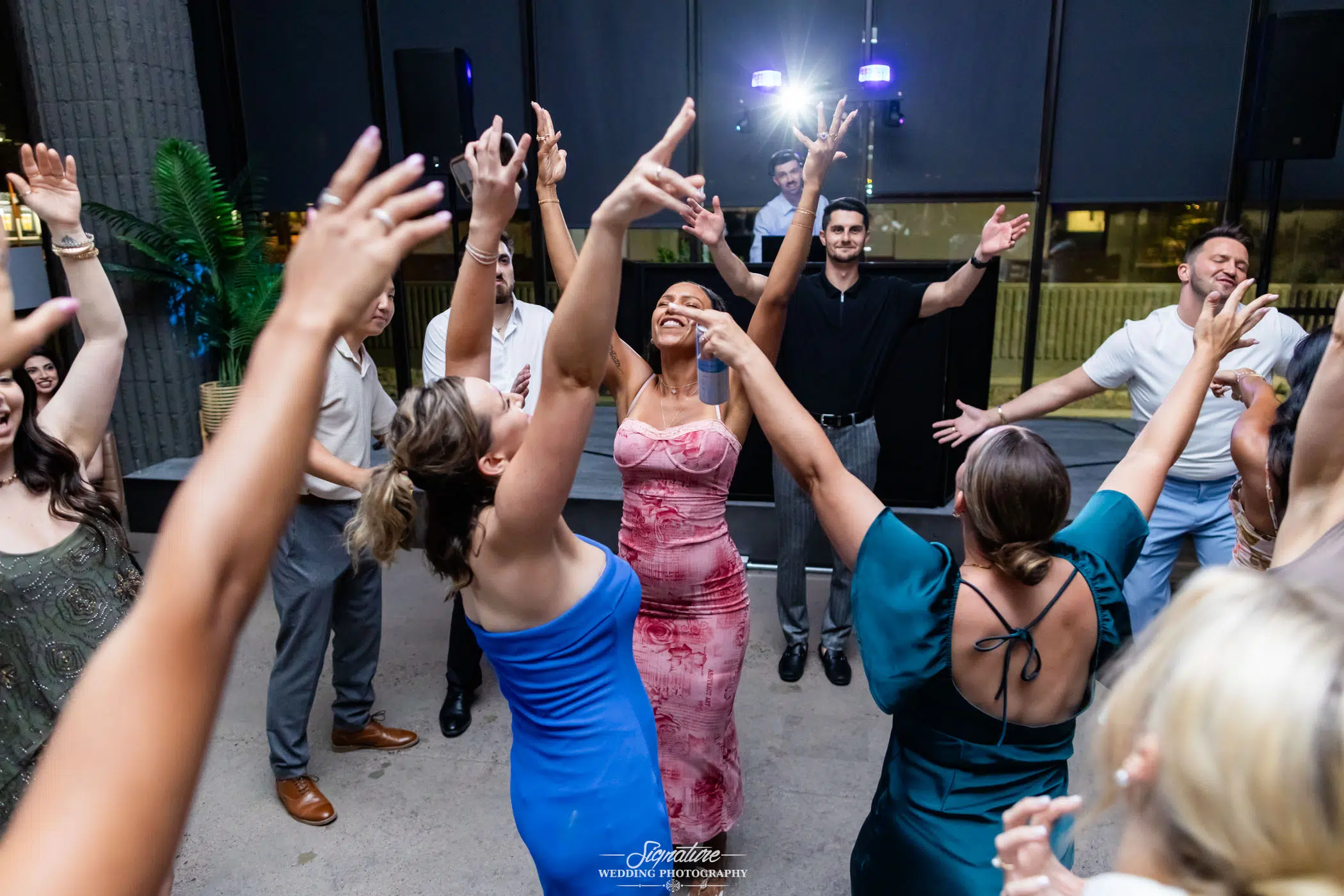 Photo by Tavits Photography A group of people enthusiastically dance in a circle at an indoor party, their arms raised and smiles on their faces, with colorful dresses and suits, under bright lights.