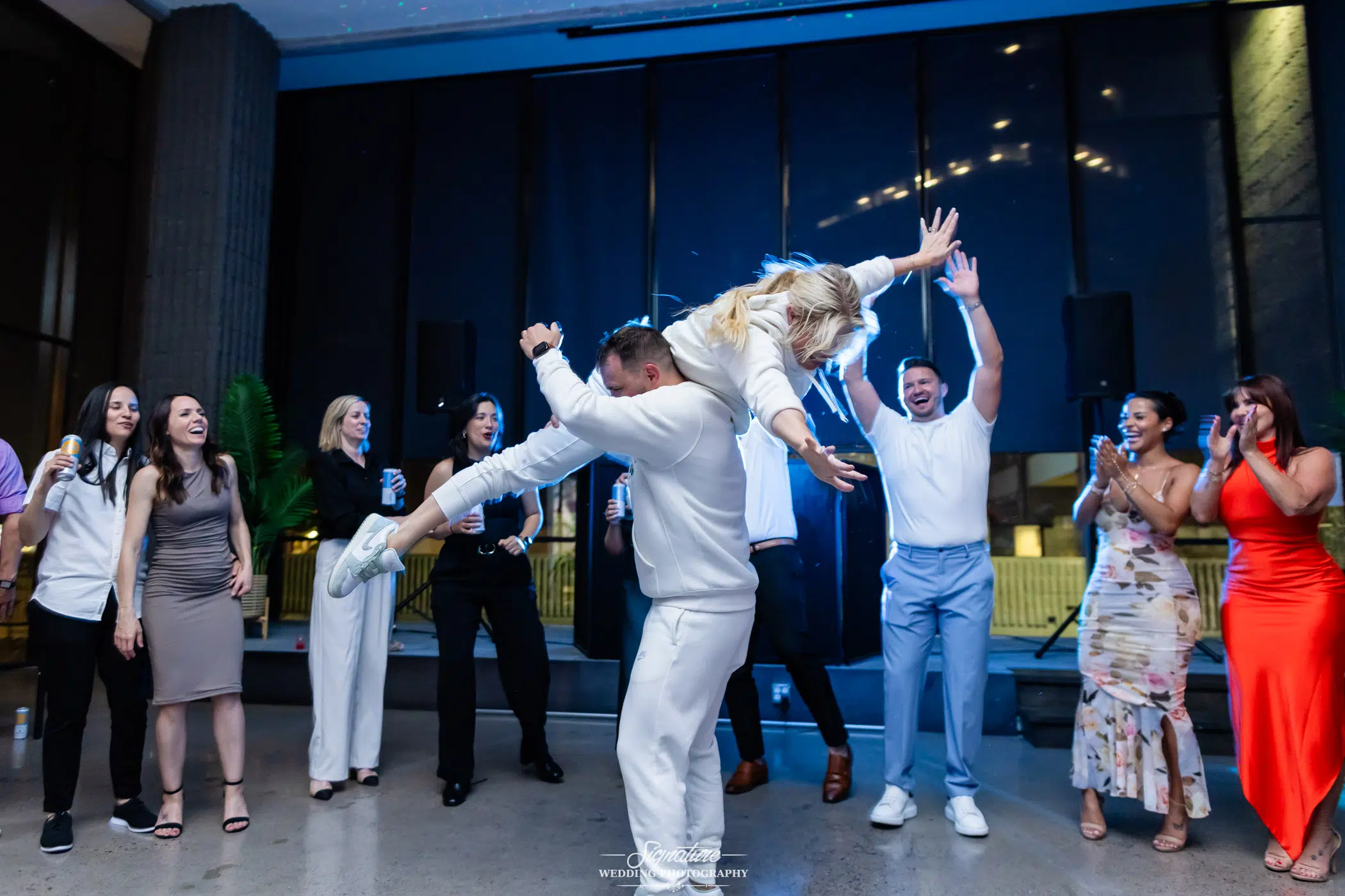 Photo by Tavits Photography A man lifts a woman in the air while dancing at a party, surrounded by smiling and cheering friends dressed in semi-formal attire. The room has large windows and a lively atmosphere.