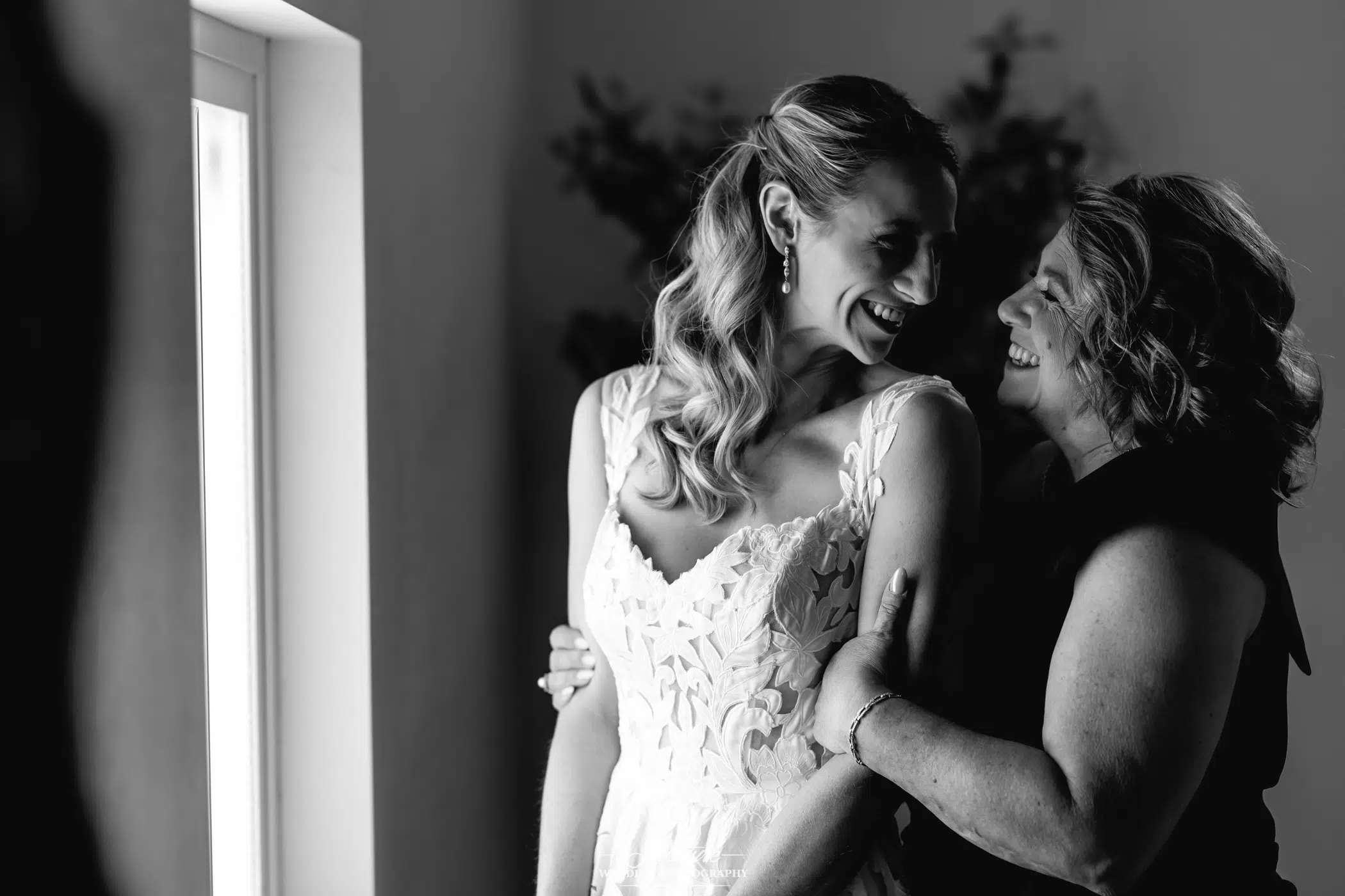 A bride in a lace wedding dress smiles and embraces a woman, both laughing together near a window with soft natural light. The moment is joyful and intimate, captured in black and white.