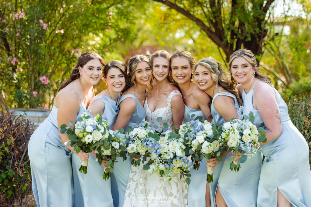 A bride in a white dress and six bridesmaids in pale blue dresses smile and pose together outdoors, each holding bouquets of white and blue flowers, with greenery and sunlight in the background.
