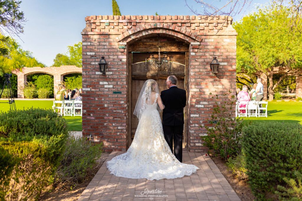 A bride in a white gown and veil stands arm-in-arm with an older man in a suit, facing a rustic brick archway outdoors, with guests seated on white chairs on a sunny lawn ahead.