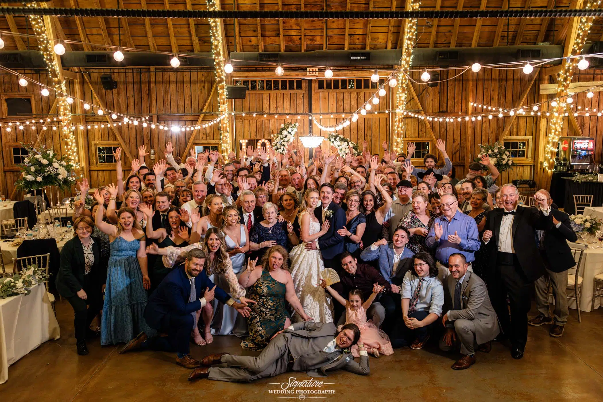 Photo by Tavits Photography A large wedding group poses for a joyful photo inside a rustic barn decorated with string lights. The bride and groom stand in the center, surrounded by smiling guests, many raising their hands and celebrating.