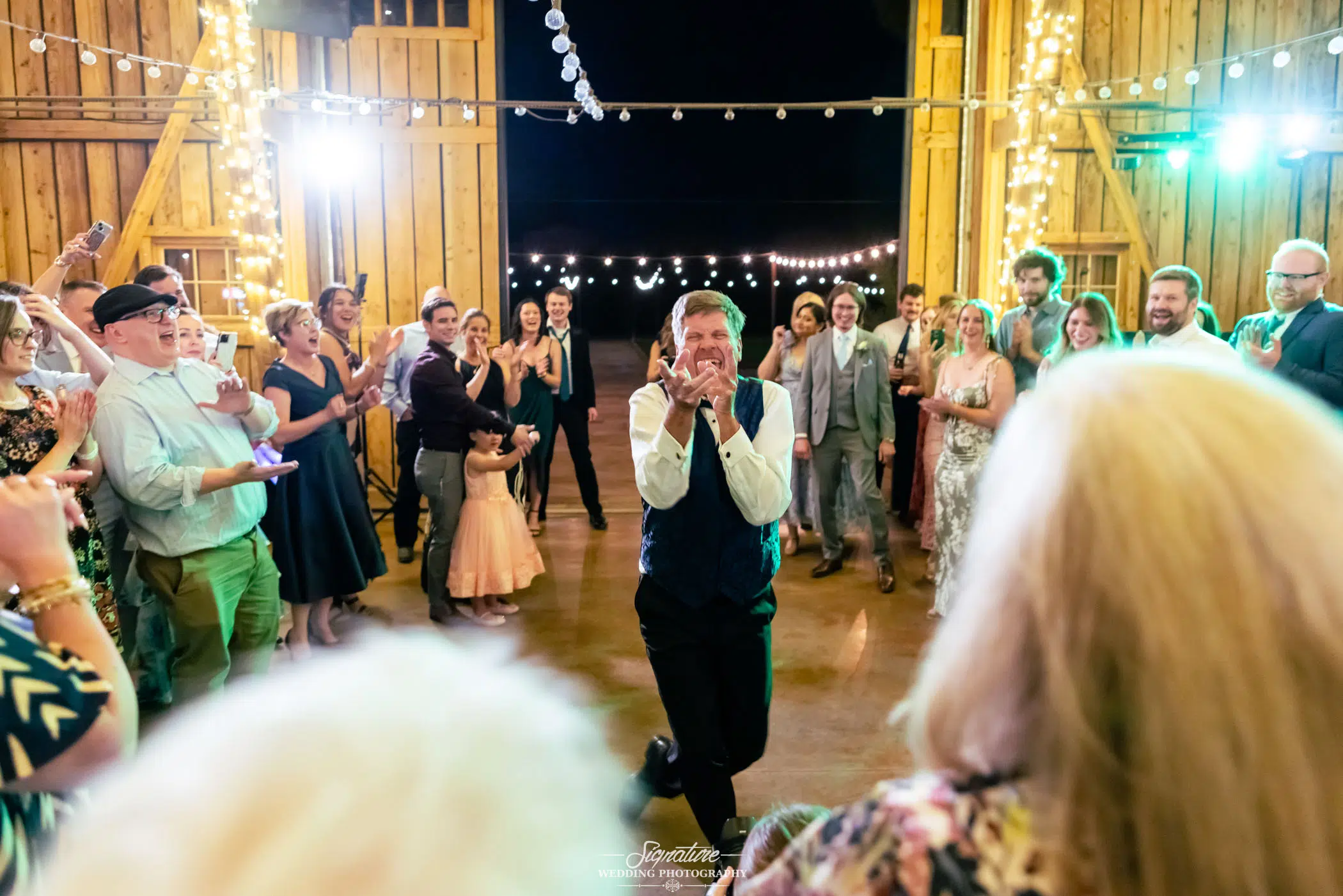Photo by Tavits Photography A joyful man in a vest stands in the center of a barn, clapping his hands to his face while smiling. He is surrounded by a cheering crowd under string lights, with people dressed for a festive celebration.