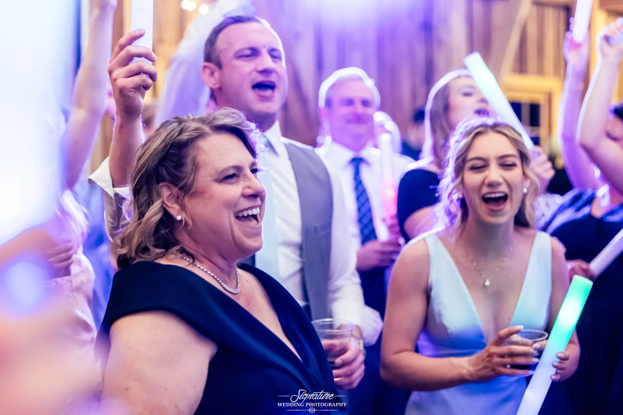 Photo by Tavits Photography A group of people, dressed formally, are laughing and cheering at a lively indoor event, holding drinks and glowing sticks, with bright lights creating a festive atmosphere.