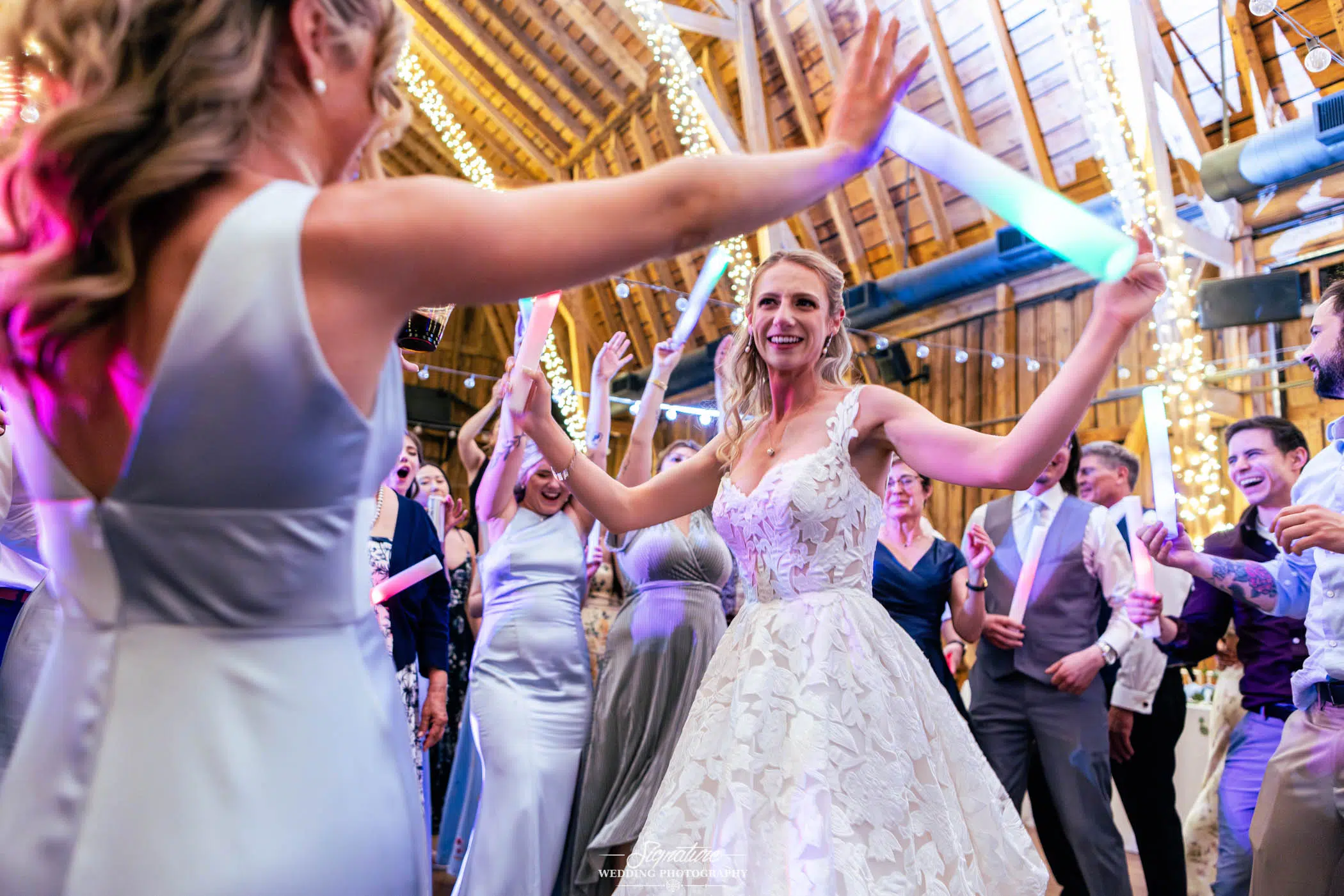 Photo by Tavits Photography A smiling bride in a white gown dances energetically with guests at a lively wedding reception in a barn decorated with string lights. People around her are cheering and holding glowing sticks.