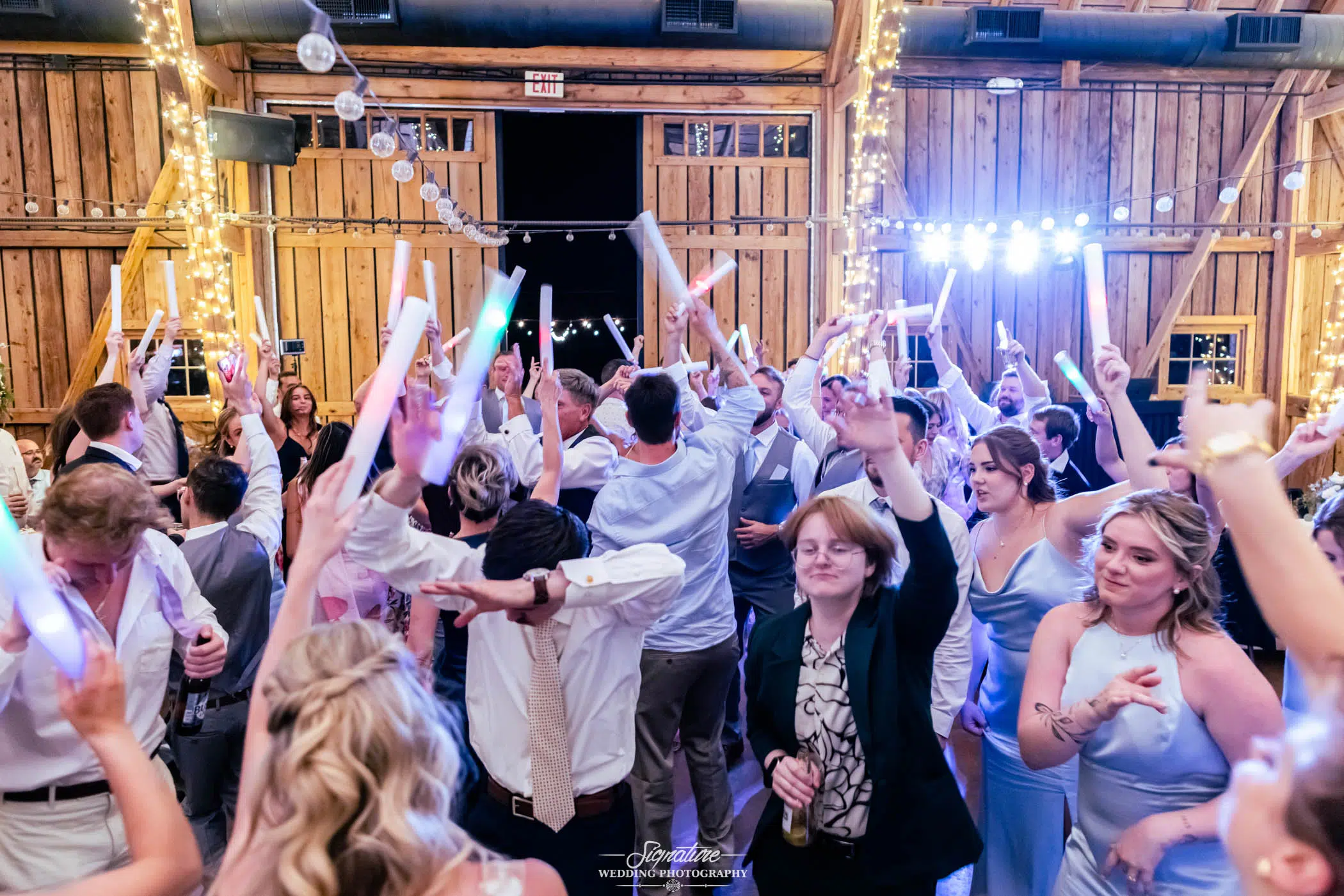 Photo by Tavits Photography A lively group of people dance and celebrate under string lights in a wooden barn, many holding glowing sticks in the air. The atmosphere is festive, with guests dressed in semi-formal attire.
