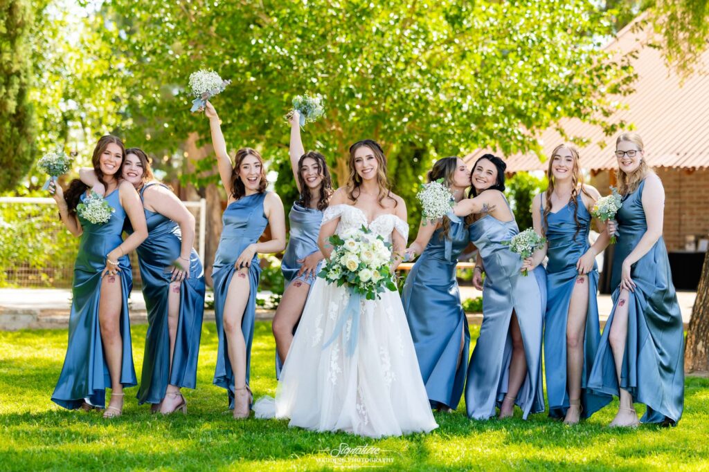 A bride in a white gown stands smiling with eight bridesmaids in shiny blue dresses, all holding bouquets and posing playfully together outdoors on a sunny day with green trees in the background.