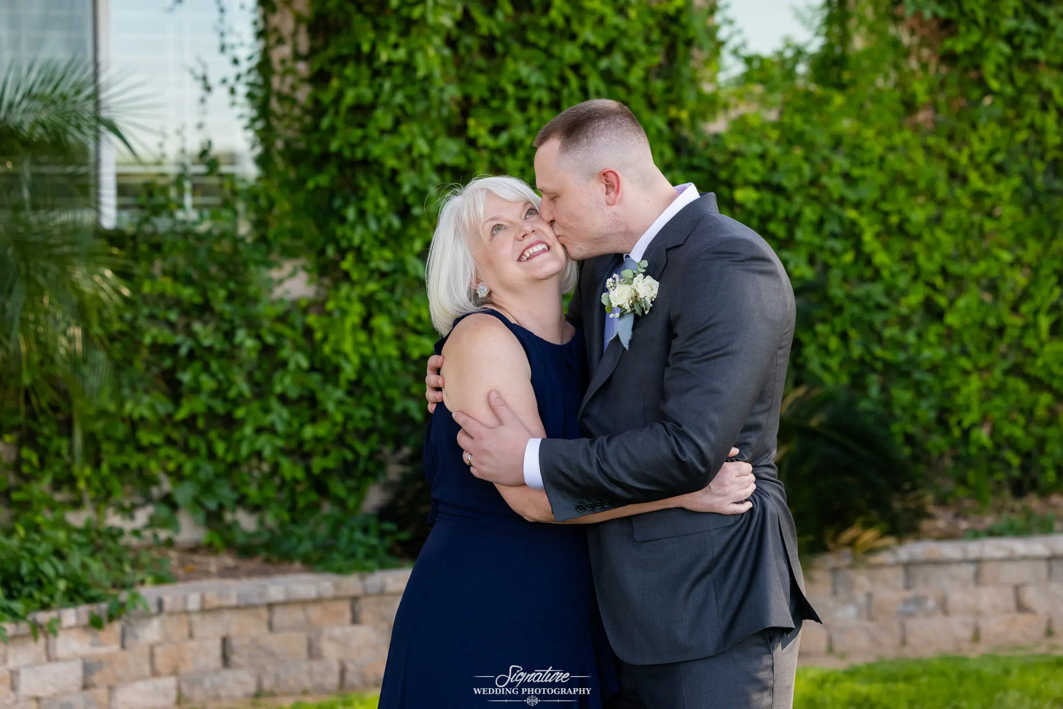 Image by Signature Wedding Photography A man in a suit hugs and kisses an older woman on the cheek outdoors. Both are smiling warmly and standing in front of a green leafy background and a low stone wall.