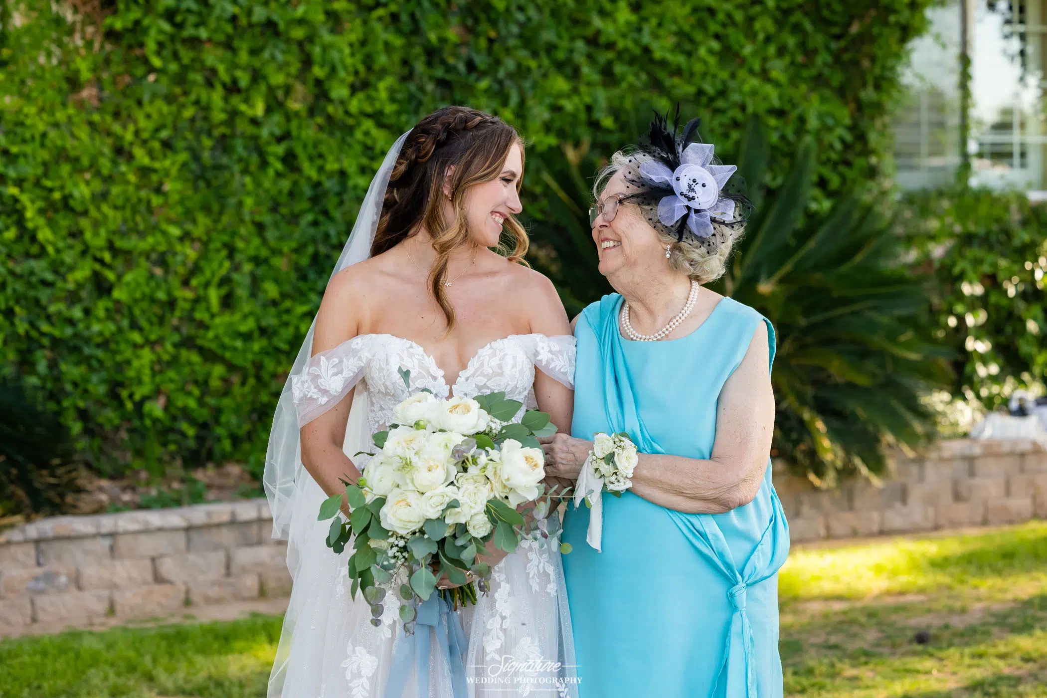 Image by Signature Wedding Photography A bride in a white dress holding a bouquet smiles warmly at an older woman in a light blue dress and floral headpiece; they stand outdoors in front of greenery.