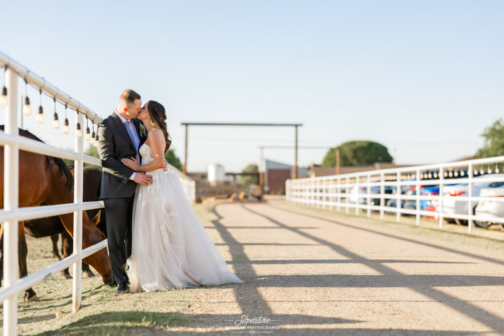 A bride and groom stand close together on a ranch pathway, leaning against a white fence. Horses graze nearby, and parked cars are visible in the background under a clear, sunny sky.